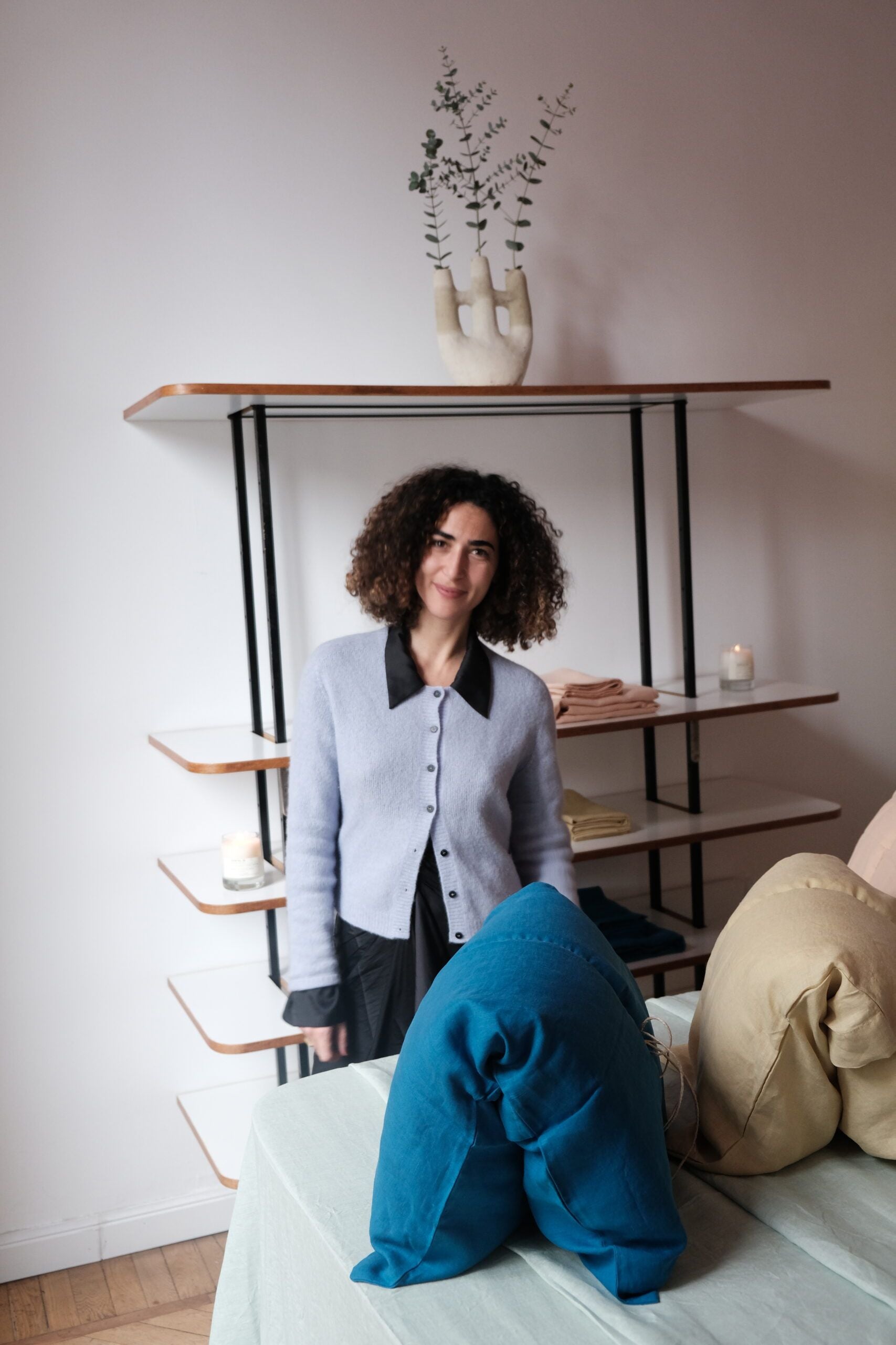 A woman with curly hair stands in front of a shelf with folded towels, a candle, and a vase of greenery. In the foreground are neatly stacked blue and beige cushions.