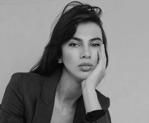 Black and white photo of a woman with long dark hair, wearing a blazer, resting her face on her hand, and looking directly at the camera with a neutral expression.