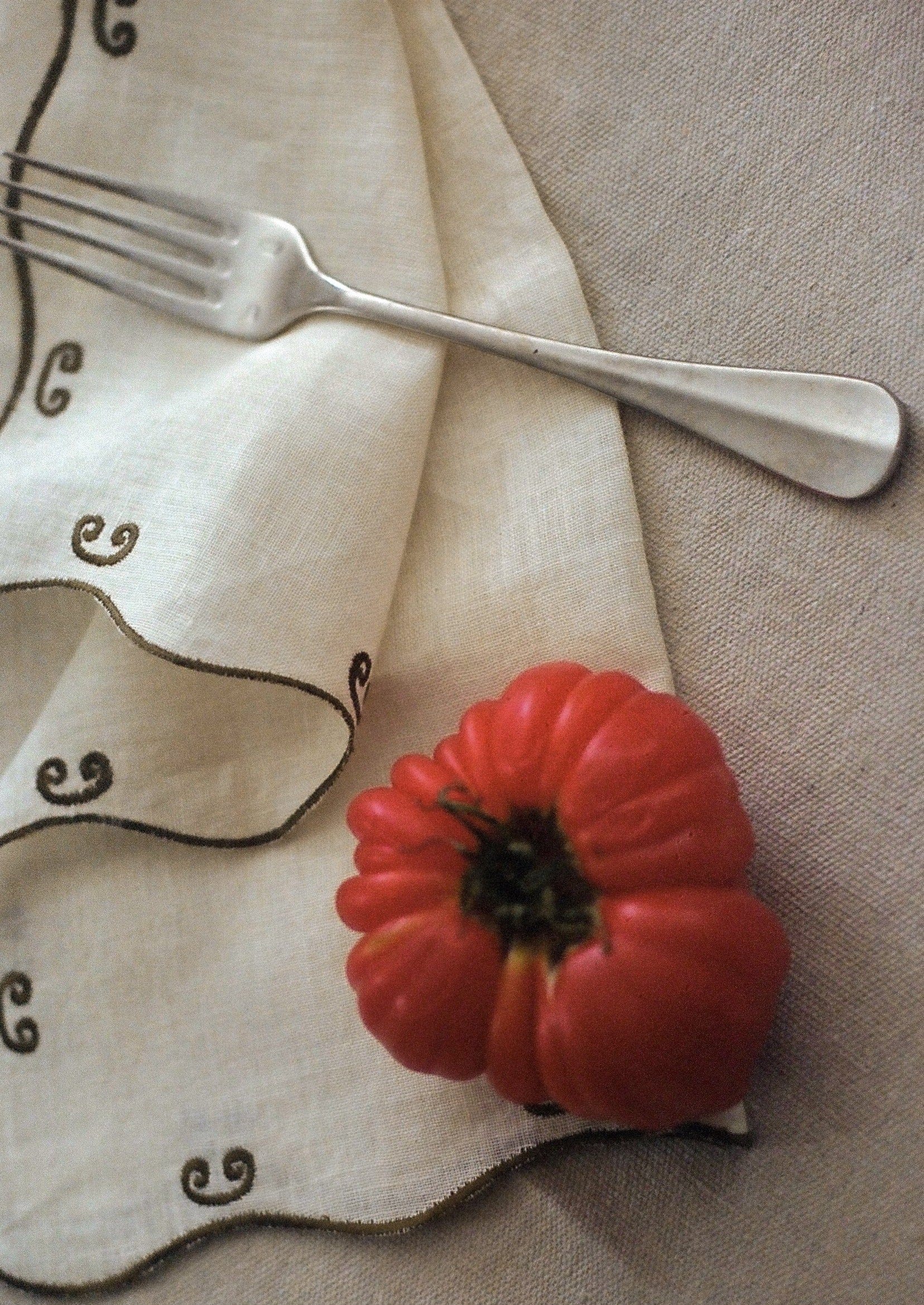 A large, red, heirloom tomato rests on a beige cloth with curly embroidery next to a silver fork on a light brown tablecloth.