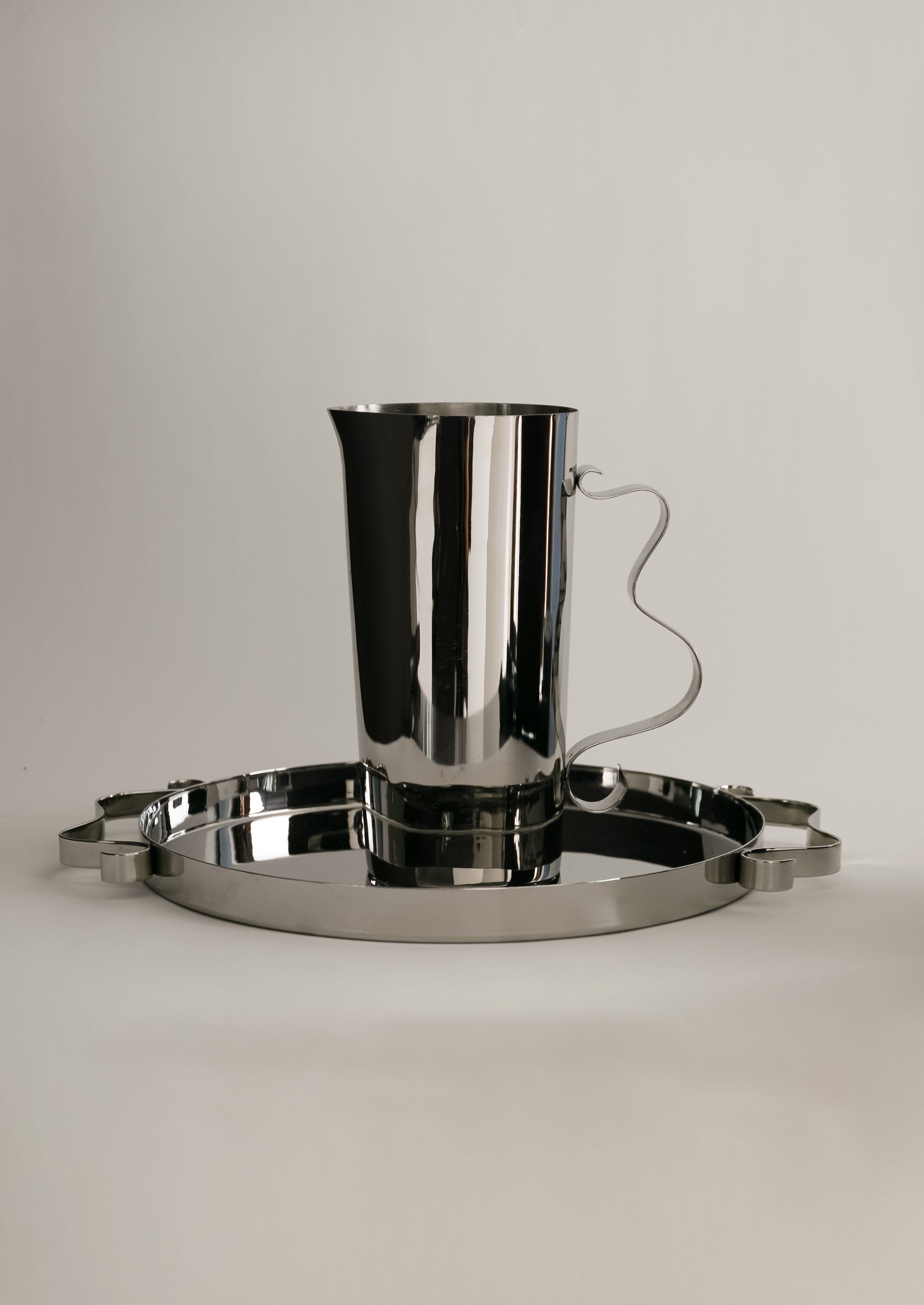 A polished silver pitcher with a sleek, curvy handle sits on an oval silver tray with small feet. The background is plain, allowing the reflective surfaces to stand out.