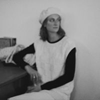 Black and white photo of a person sitting beside a table, wearing a light beret and a sleeveless sweater over a dark long-sleeve shirt, looking off to the side. The background is plain.