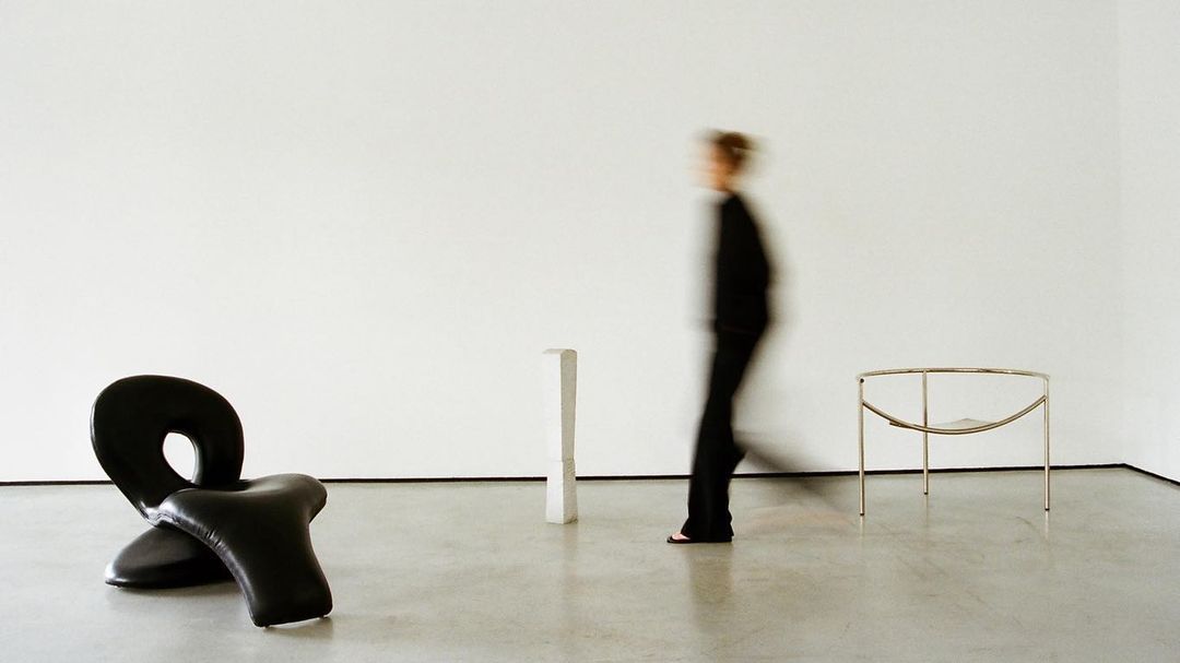 A blurred person walks past a modern black chair, a small white pedestal, and a metal-framed table in a minimalist room with a plain white wall.
