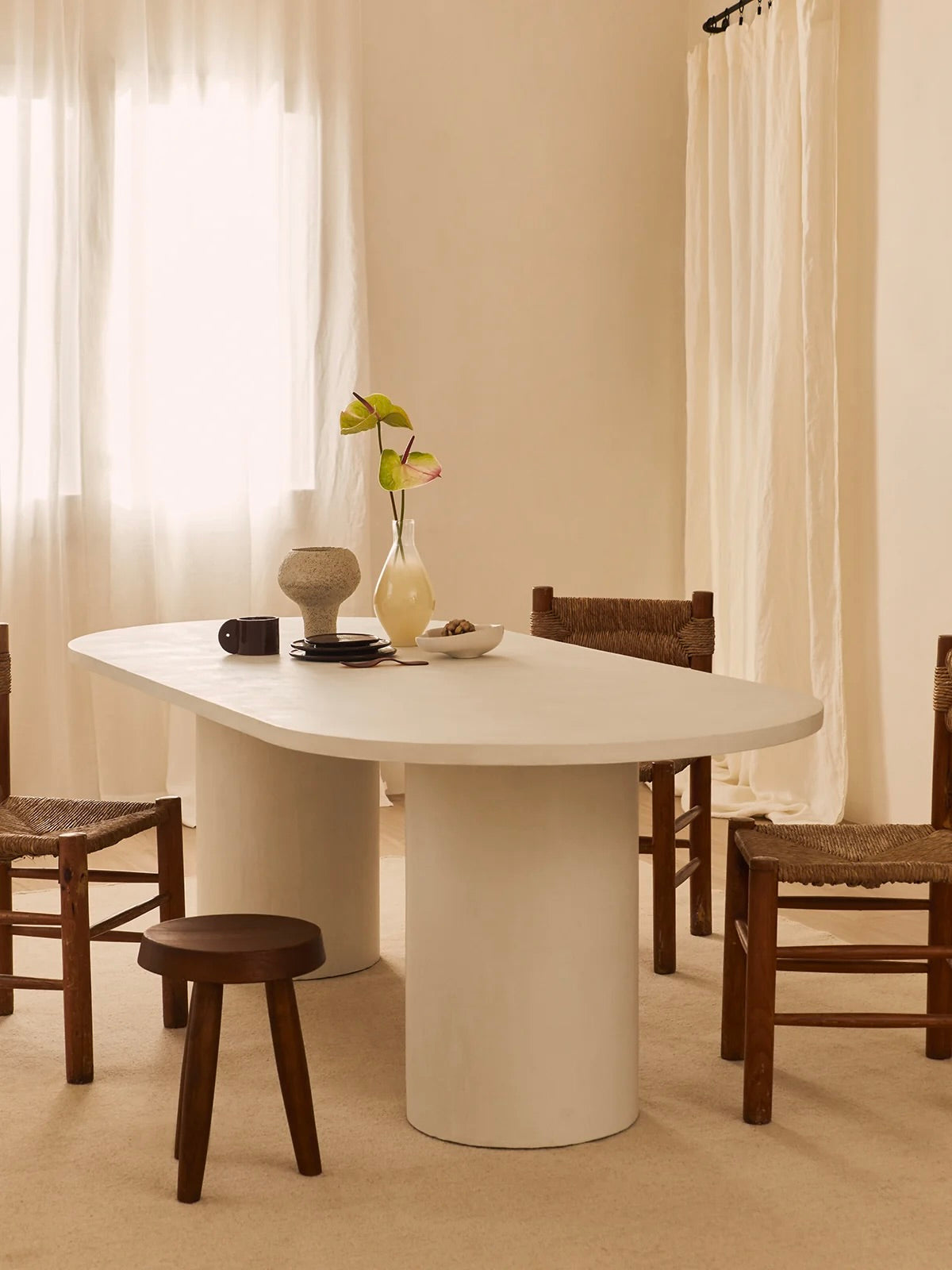 A minimalist dining room featuring a white, oval-shaped table with cylindrical legs, surrounded by wooden chairs and a small stool. On the table are a vase with flowers, a ceramic object, and a cup. Cream-colored curtains adorn the windows.