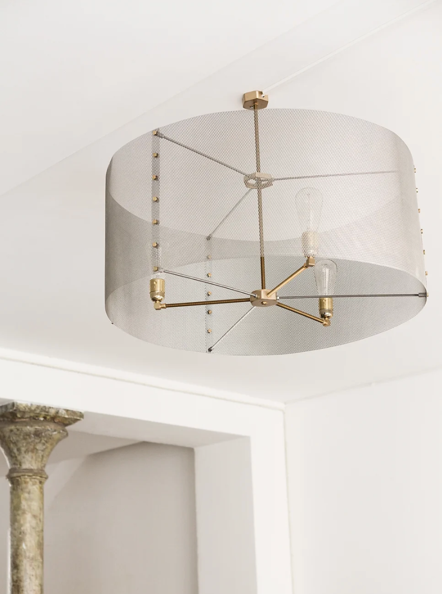 A modern pendant lighting ceiling light fixture with a cylindrical metal mesh shade and an exposed bulb design is mounted on a white ceiling. The fixture features a brass frame. A decorative column is partially visible to the left.