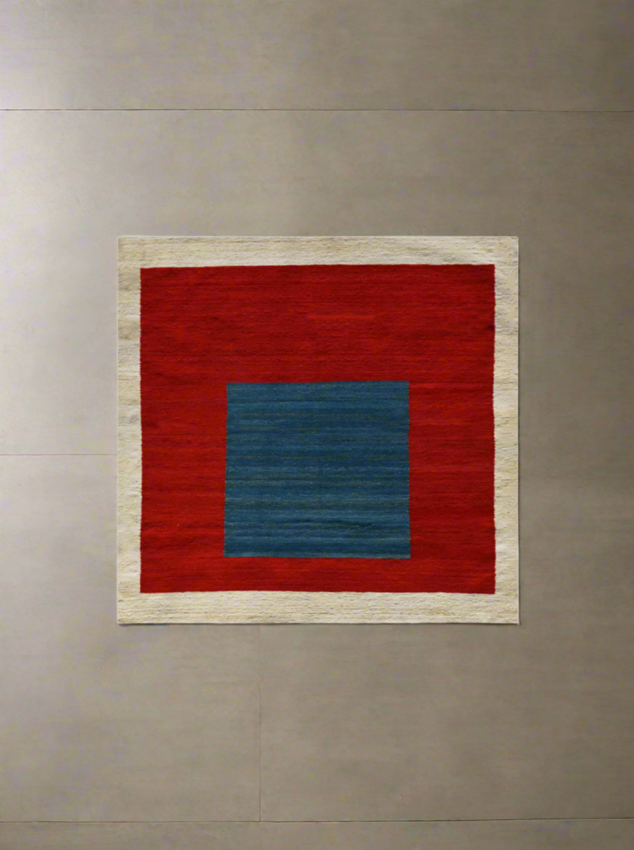 A square wall art piece featuring a large red square with a smaller blue square at the center, bordered by a thin beige frame, displayed on a plain wall.