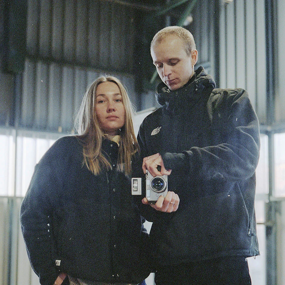 A modern, metallic candle holder with an abstract design sits on a white pedestal against a plain background. The holder supports two white, unlit candles, with its sleek, twisted form adding a contemporary touch.