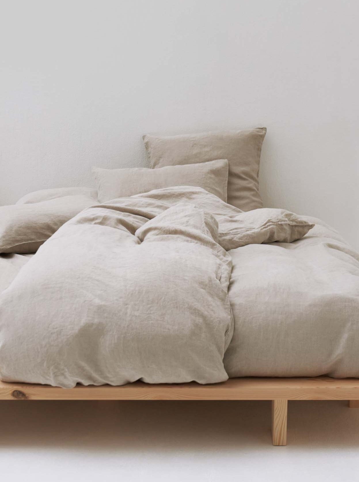 Minimalist bedroom setup with a natural wood frame bed. The bed is made up with beige linen bedding, including two pillows, one cushion, and a duvet, all arranged neatly against a plain white wall.