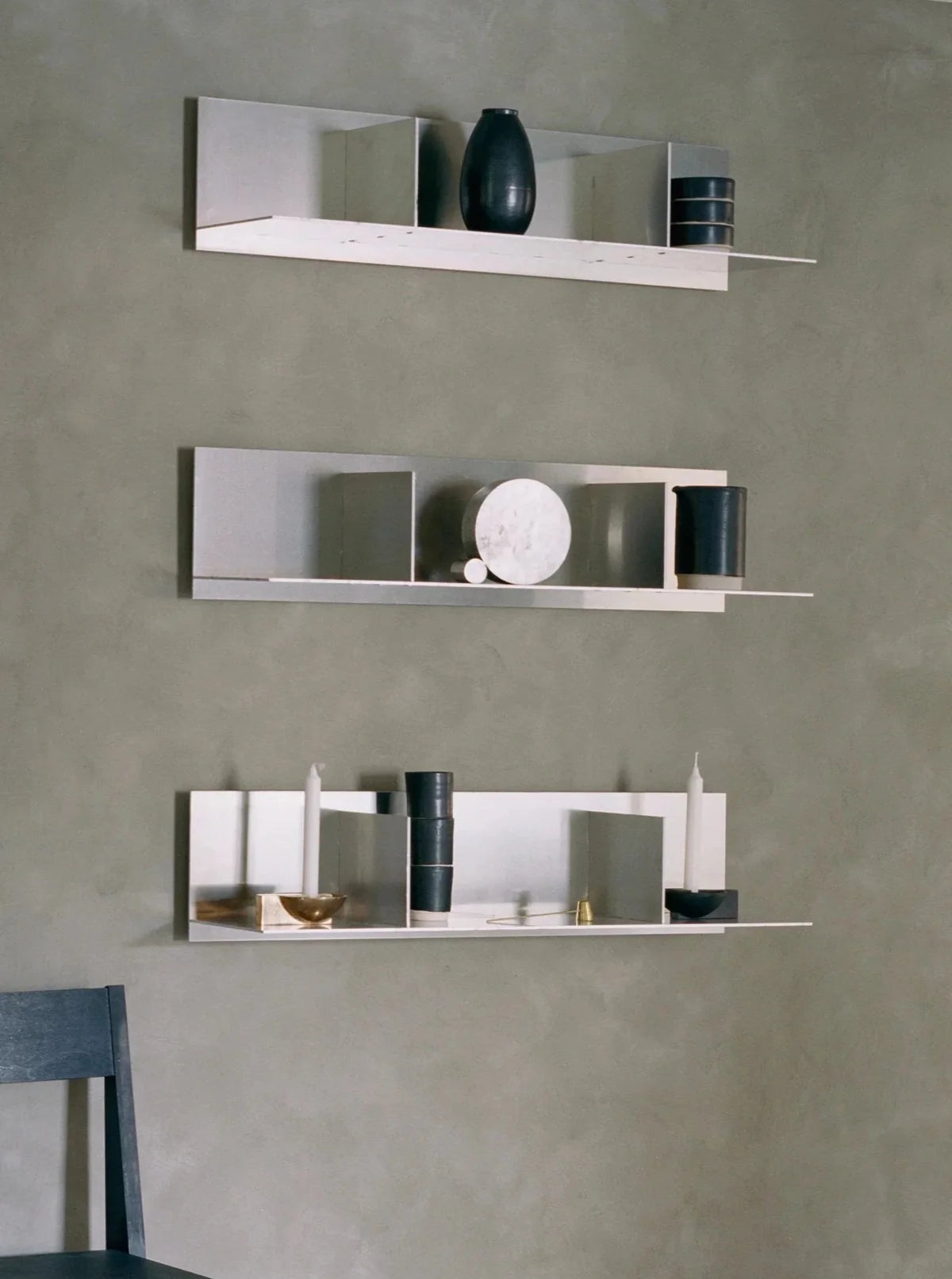 Three minimalist white shelves mounted on a grey wall, each holding a mix of decorative items including black vases, white sculptures, and books. A black chair is partially visible in the lower left corner.
