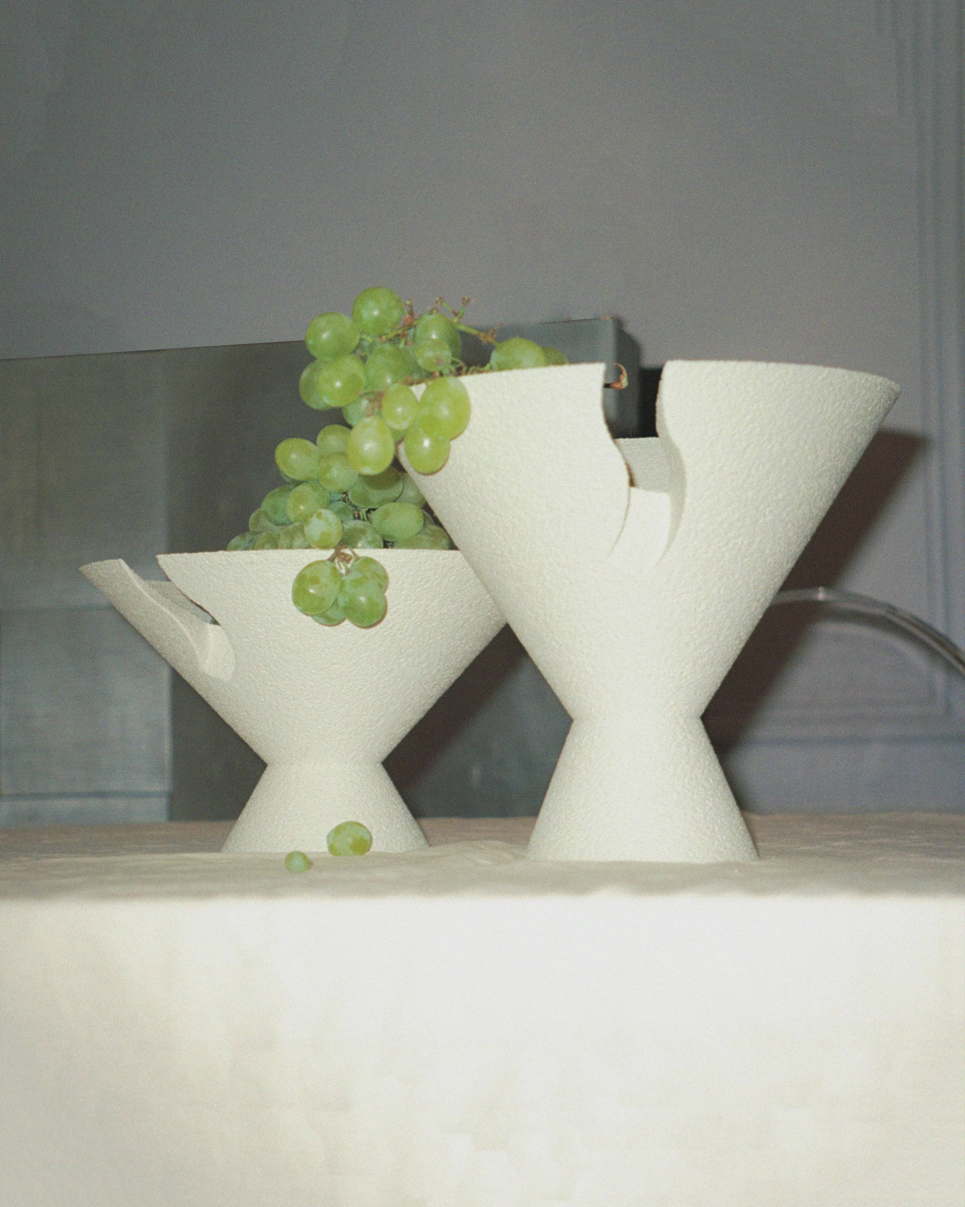 Two uniquely shaped white vases on a counter, one adorned with a bunch of green grapes, in a softly lit kitchen setting.