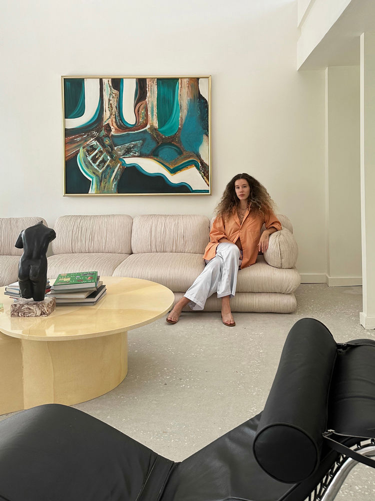 A woman with long curly hair sits on a beige sofa in a modern living room with neutral tones, abstract wall art, a round coffee table, and a black sculptural chair in the foreground.