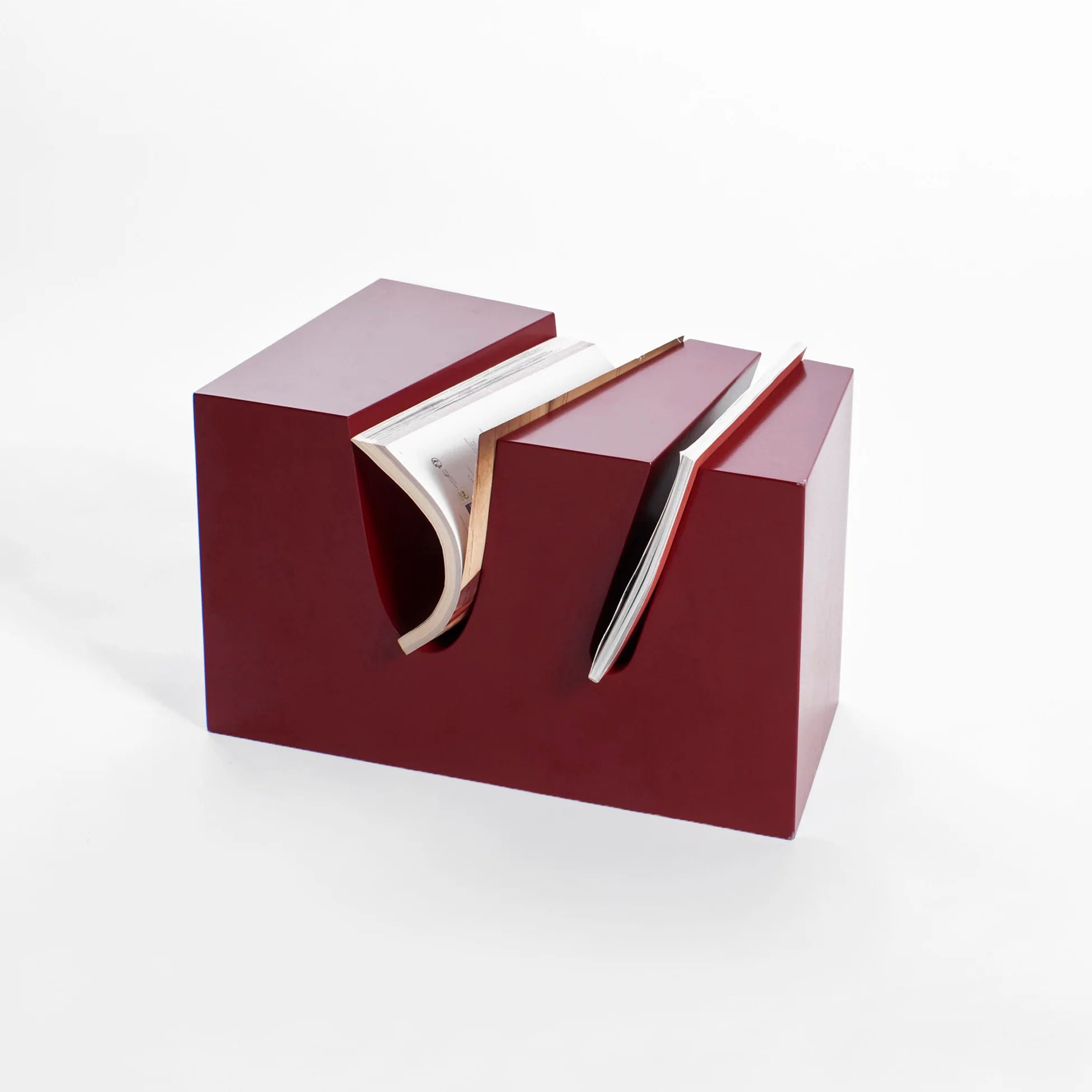 A modern, abstract book organizer made of red material with three deep grooves to hold books. The grooves create an angular, wave-like pattern, giving the piece a contemporary and artistic appearance. One book is partially placed in the center groove.