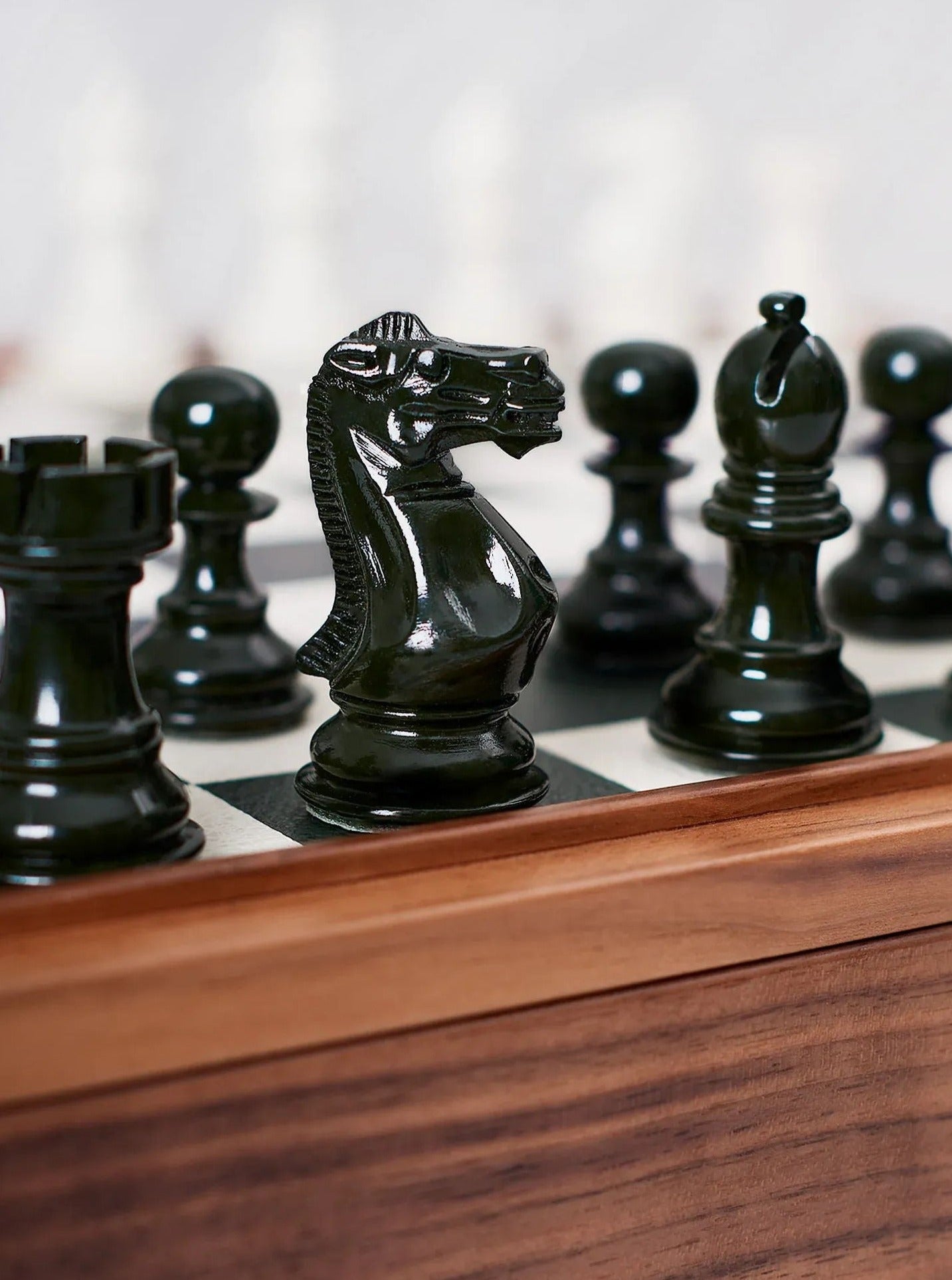 A close-up of black chess pieces on a chessboard, with focus on a knight in the foreground. The wooden chessboard has a light and dark square pattern. The pieces are glossy and detailed.