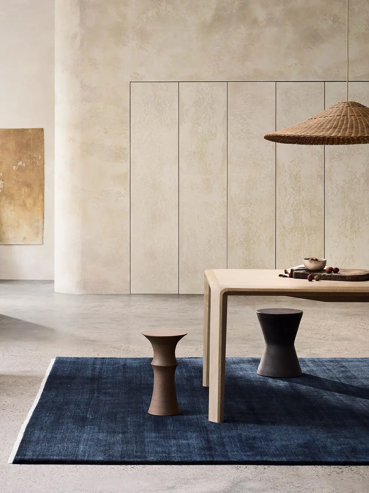A minimalist living space featuring a light wooden bench on a deep blue rug, two dark ceramic stools, and a wicker lamp overhead. A textured beige wall and abstract art piece complete the serene ambiance.