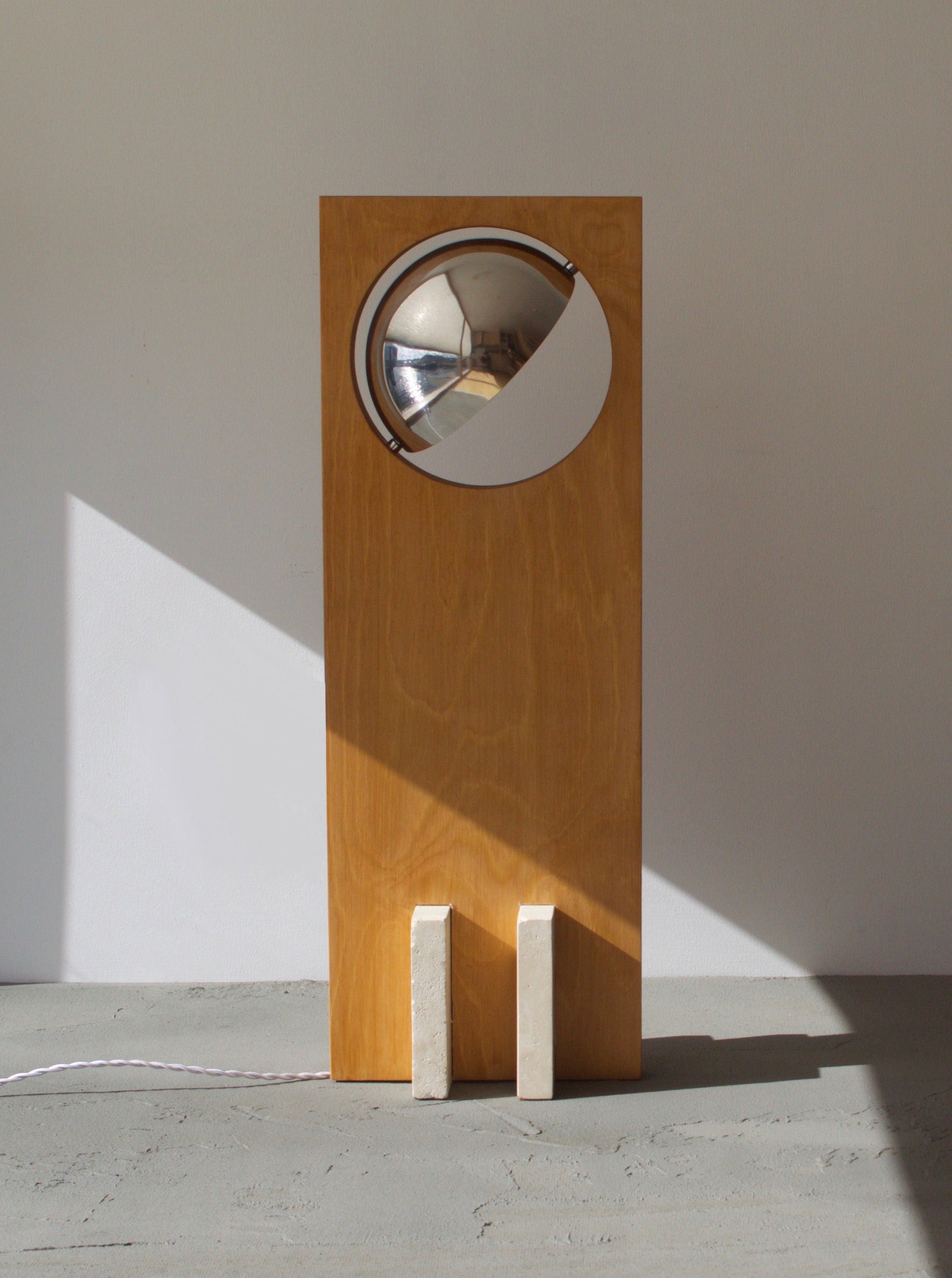 A modern art floor lamp named Galileo 2.3 floor lamp from Edoardo Lietti Studio with a circular mirror and a visible bulb, casting a sharp geometric shadow on a plain wall in a sunlit room.