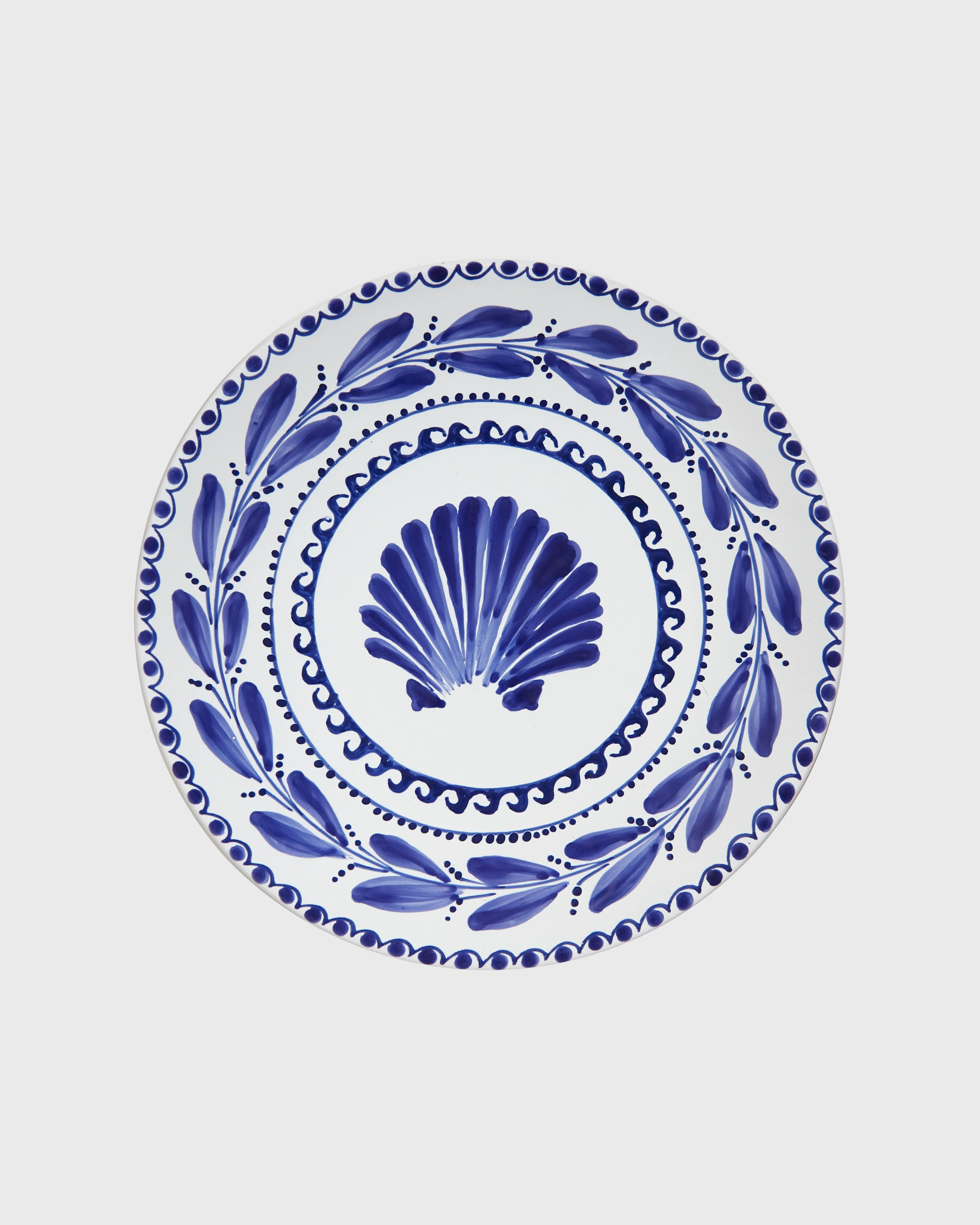 White ceramic plate with intricate blue painted design featuring a central scallop shell motif and decorative leaf patterns around the border, set against a plain background