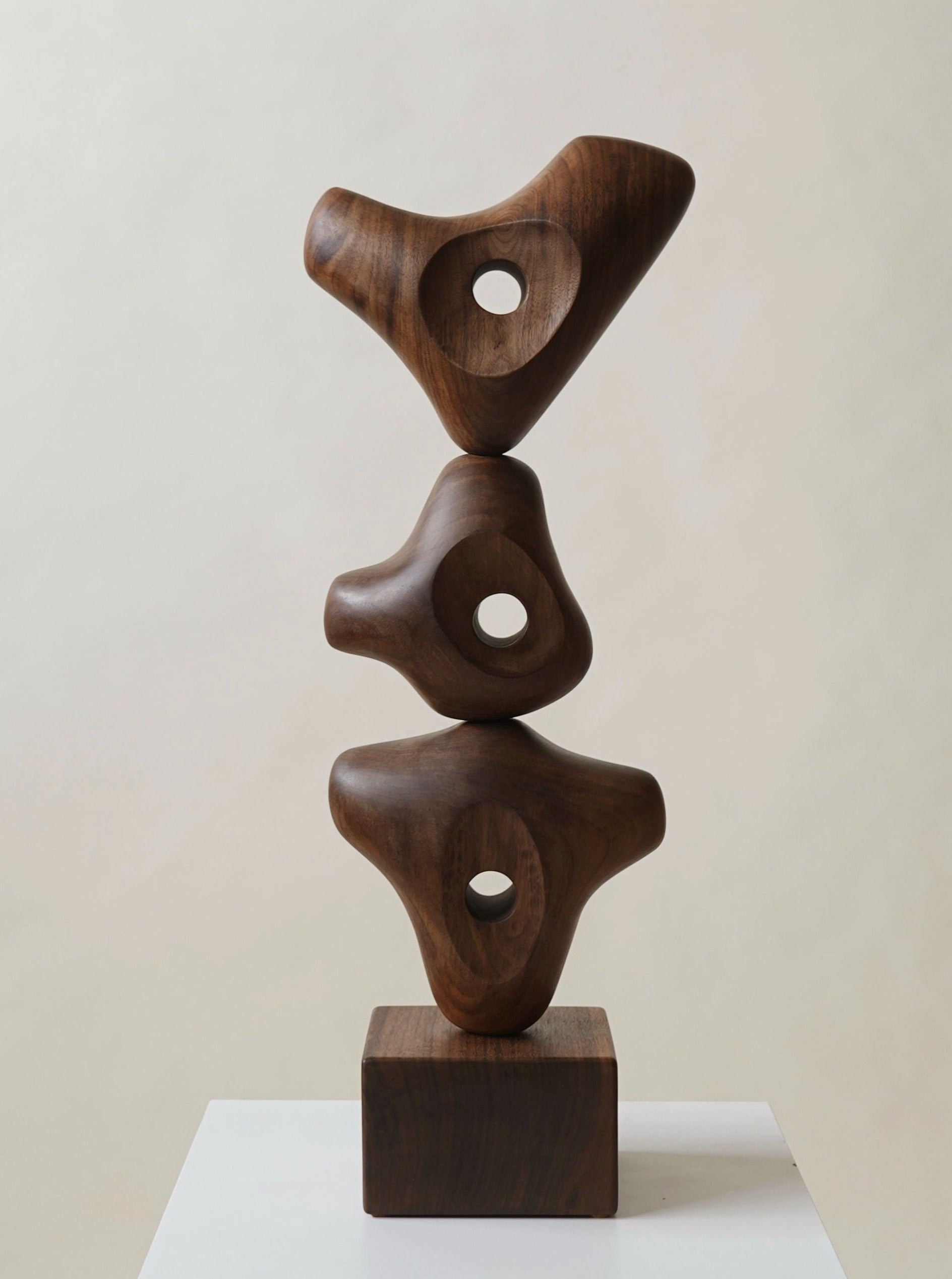 A wooden sculpture featuring three abstract forms, each with a smooth, curved shape and central hole, stacked vertically on a wooden base. The sculpture has a natural, polished finish and is displayed against a plain background.