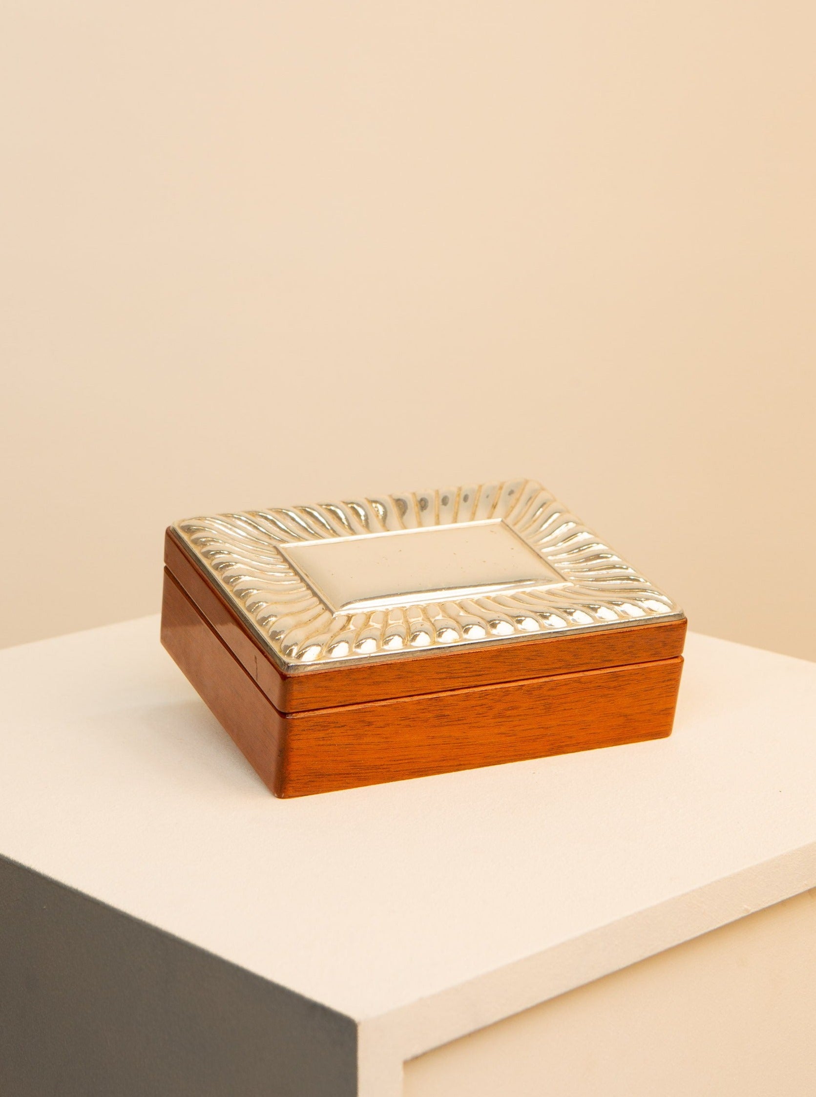 A small rectangular wooden box with an ornate, decorative silver lid sits on a two-toned pedestal. The lid features an intricate border pattern, and the overall design has a vintage aesthetic. The background is softly lit in a neutral tone.