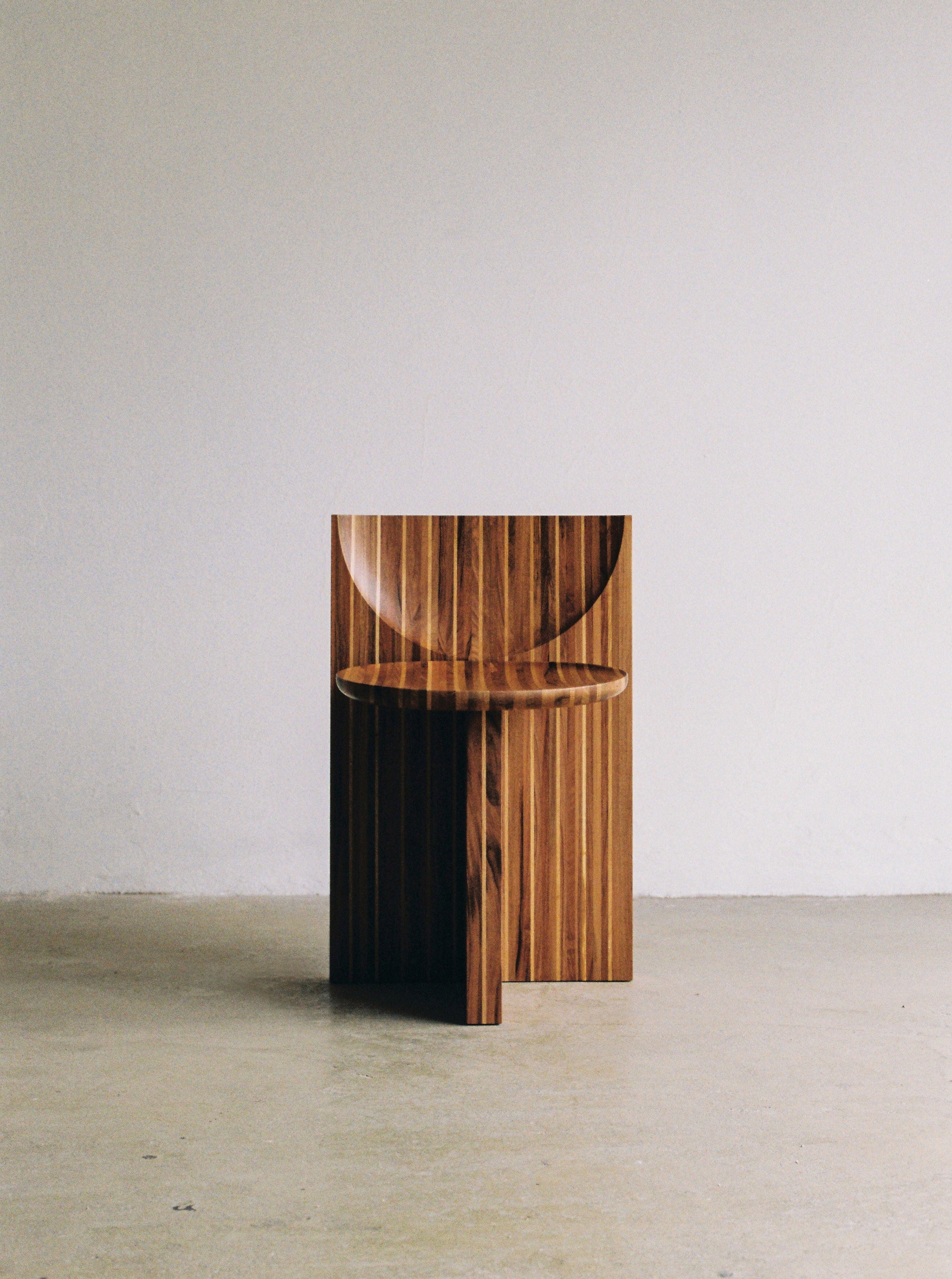 A modern wooden chair with a distinctive geometric design featuring vertical stripes, positioned against a plain, light-colored wall in a minimalist, empty room with a smooth floor.