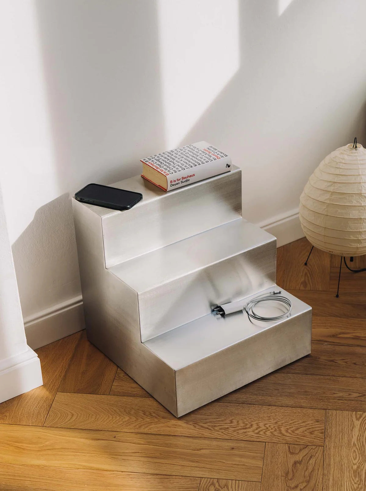 A small, three-tiered metal shelf placed against a wall, holding a smartphone, a book, and a coiled charging cable. The shelf is on a wooden floor, and a paper lantern is positioned nearby. Sunlight illuminates the scene.