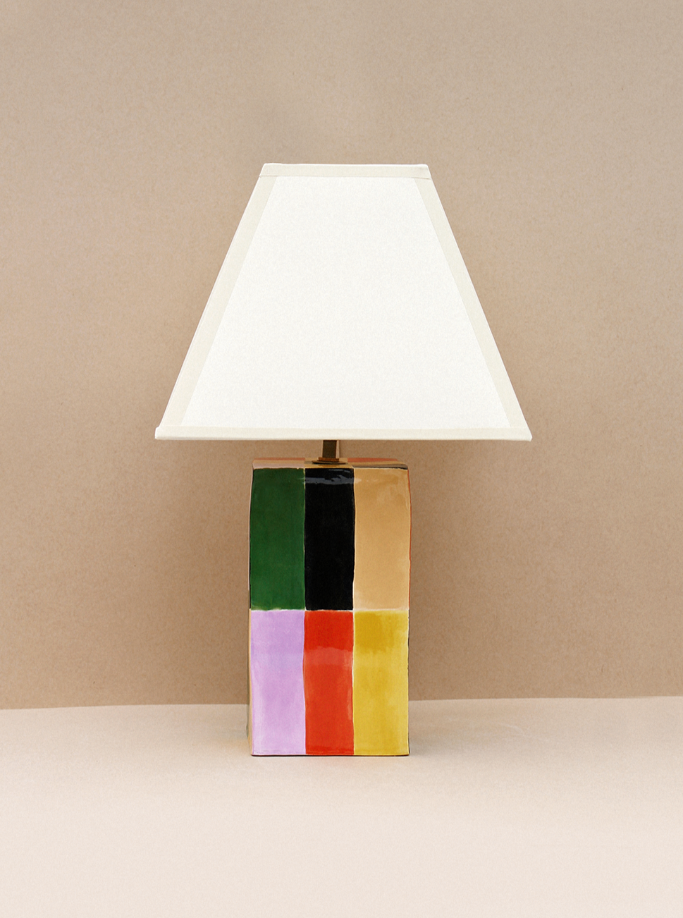 A table lamp with a white triangular lampshade sits against a beige background. The lamp base features a colorful patchwork design with squares of black, green, pink, orange, red, yellow, and beige.