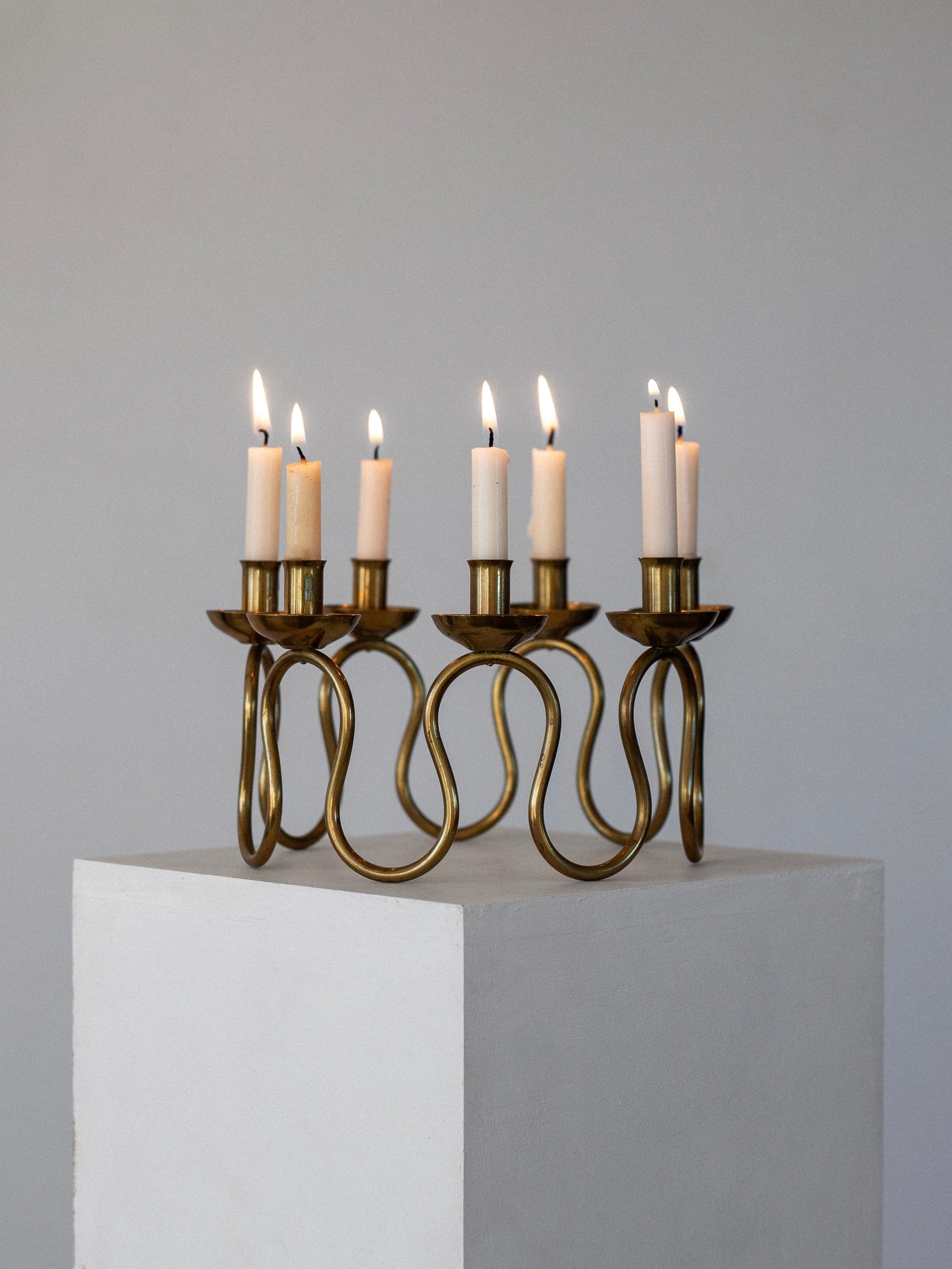 The Spigel Scandinavian Brass Candleholder from the 1940s, featuring an intricate wavy design and reminiscent of Lars Holmström's craftsmanship, is displayed with eight lit candles on a white pedestal against a neutral gray background.
