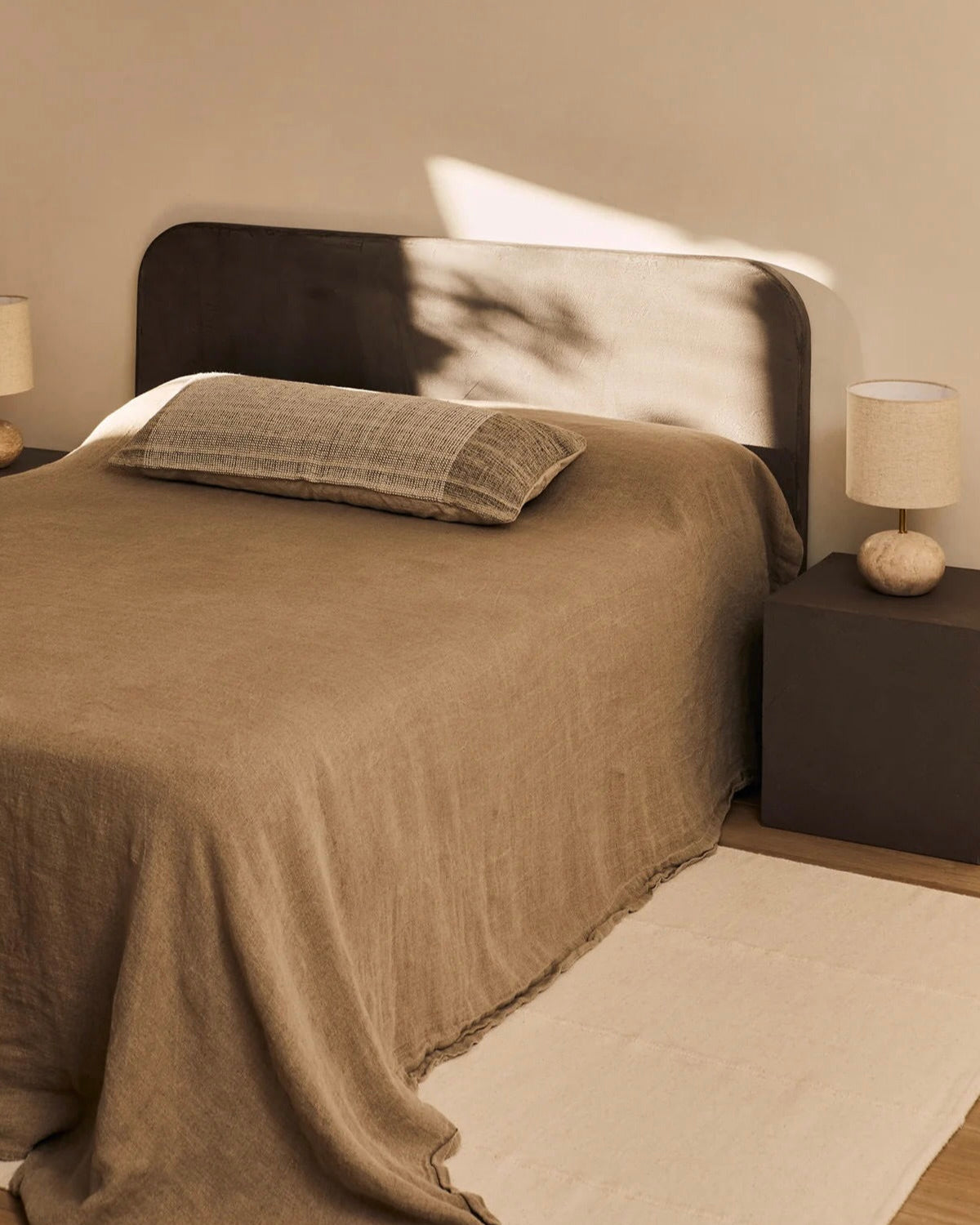 A minimalist bedroom showcases a bed with the "Ruperto" Headboard by MarlotBaus, featuring durable microcement. It's adorned with a brown linen duvet and pillow. Two bedside tables each support a lamp, casting soft shadows to create a calming atmosphere. A beige rug completes the scene on the wooden floor.