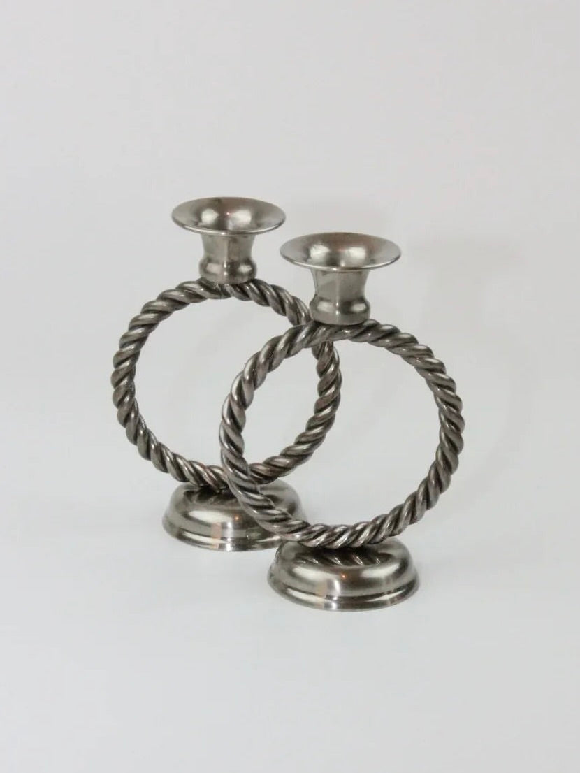 Two Mental Candle Holders by Boga Avante Shop featuring a twisted rope design forming circular holders are shown on a plain background. The bases are round and stable, with wide openings at the top to hold candles. The overall design is timeless, exuding a classic and sophisticated look that's both elegant and symmetrical.