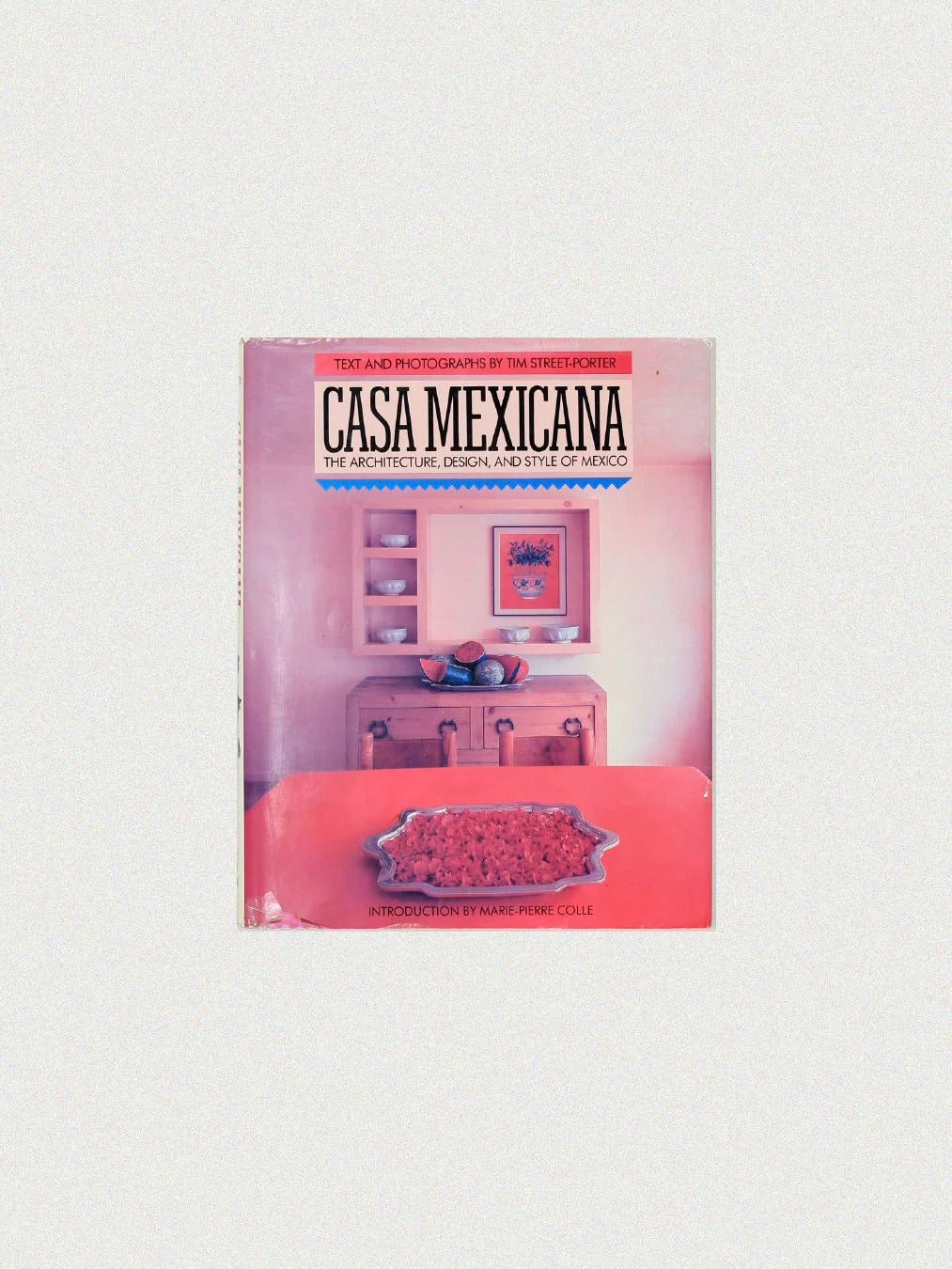 Boga Avante Shop's "Casa Mexicana" book cover elegantly highlights Mexican architecture with a room featuring pink walls, a wooden table, a pink cabinet, and a framed picture above, capturing the rich interior design and style of Mexico.