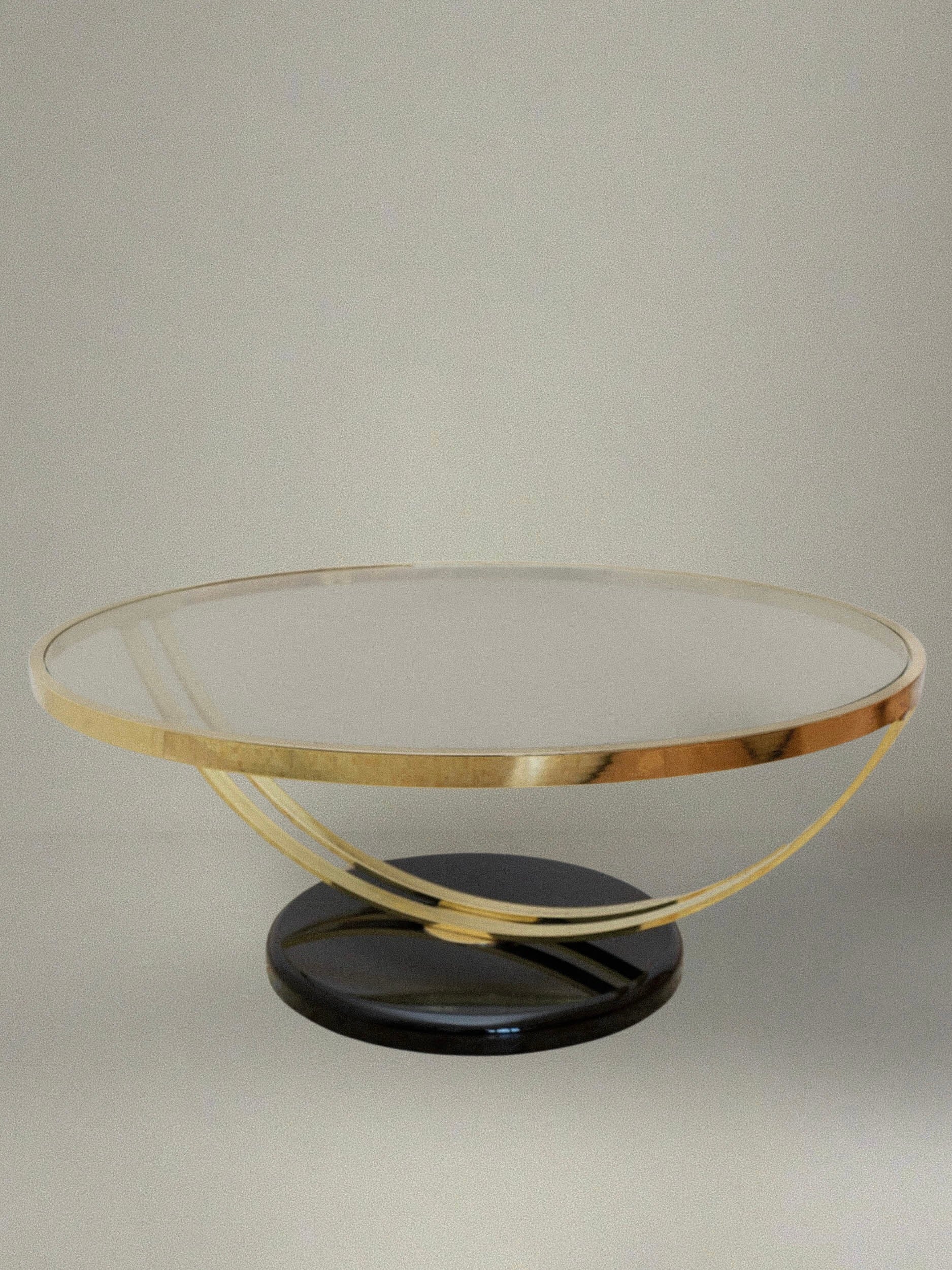 The Glass and Metal Side Table - Italy 1980s, designed by Bottega Jacobs, features a round glass top with a gold metal rim and elegantly curved supports that bring subtle Hollywood Regency charm. The sleek black circular base adds a modern touch to the minimalist design when placed against a simple, neutral-colored wall.