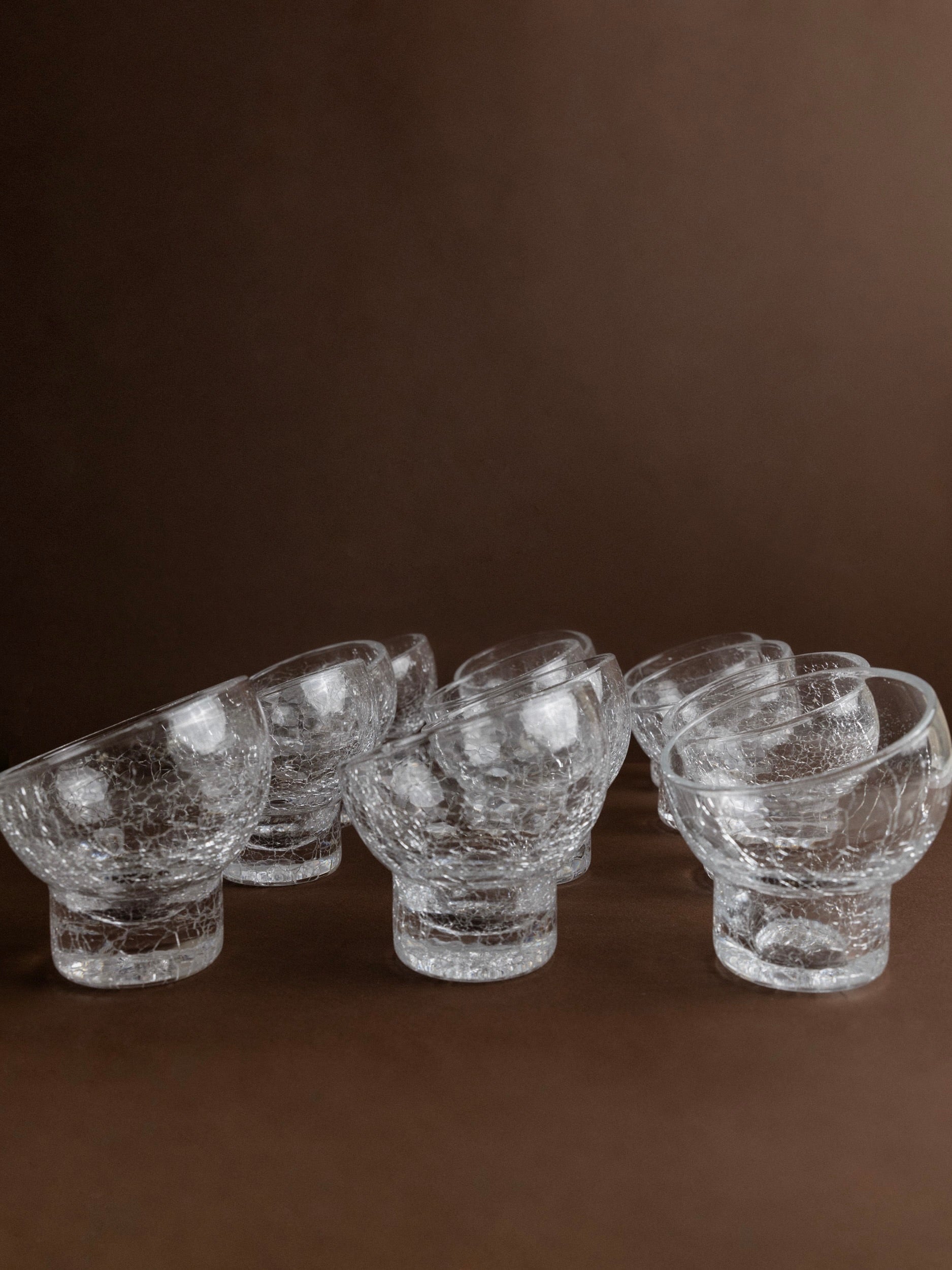 A set of 12 wine and cocktail glasses from Bottega Jacobs, Italy 1970s, is artfully arranged to form intricate patterns against a brown backdrop.