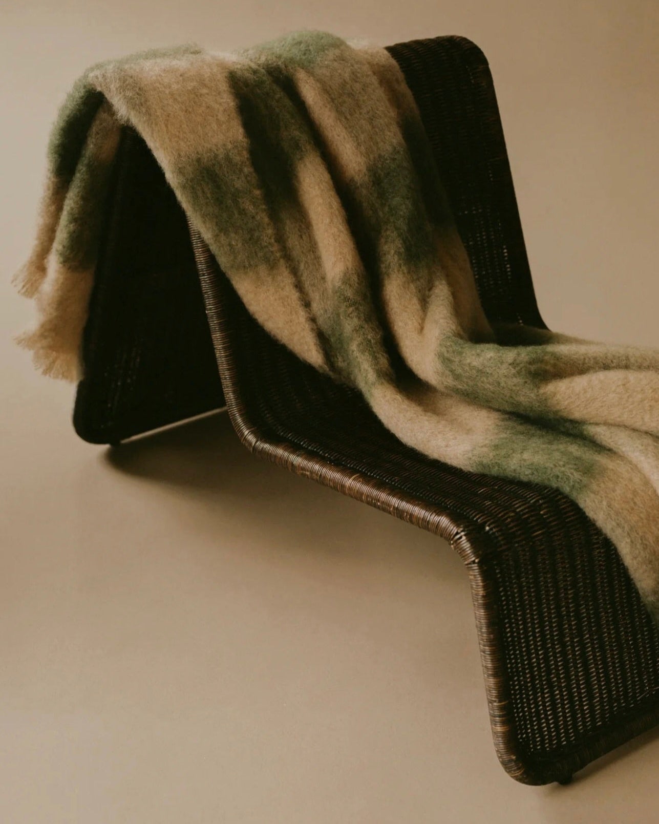 A woven, modern chair sits in a minimalist setting. Draped over it is "The Wren's" "Arbor Moss" throw, an eco-friendly, luxurious blanket in muted green and beige tones. The chair's sleek lines and the throw's texture create a cozy, stylish contrast.
