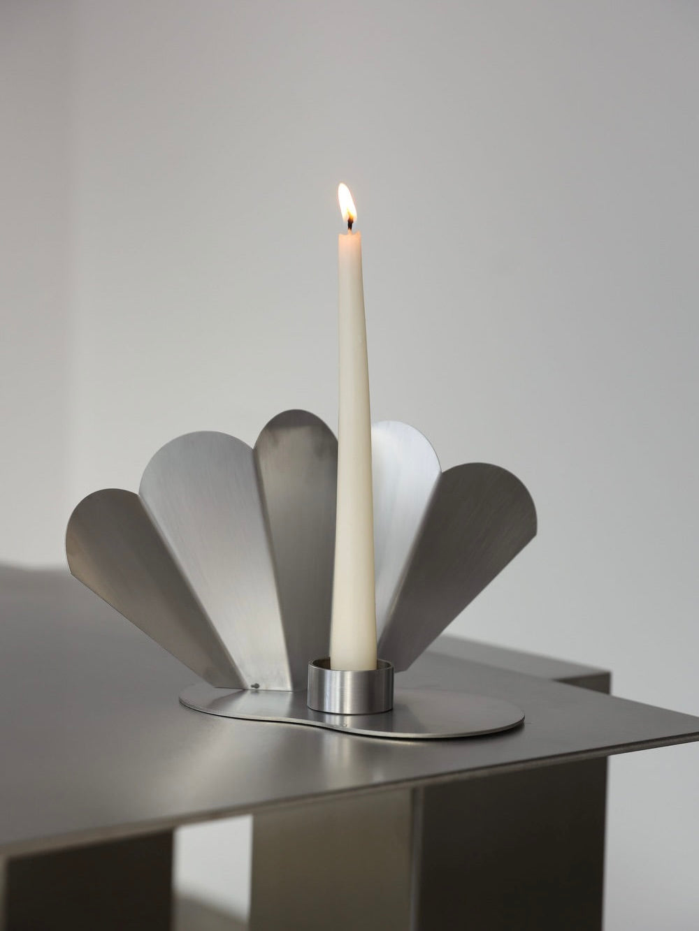 A lit white candle sits in a Paonne Candleholder by The Good Living&Co., crafted from stainless steel, on a minimalist geometric metal surface. The plain, softly lit background enhances the holder's modern design.