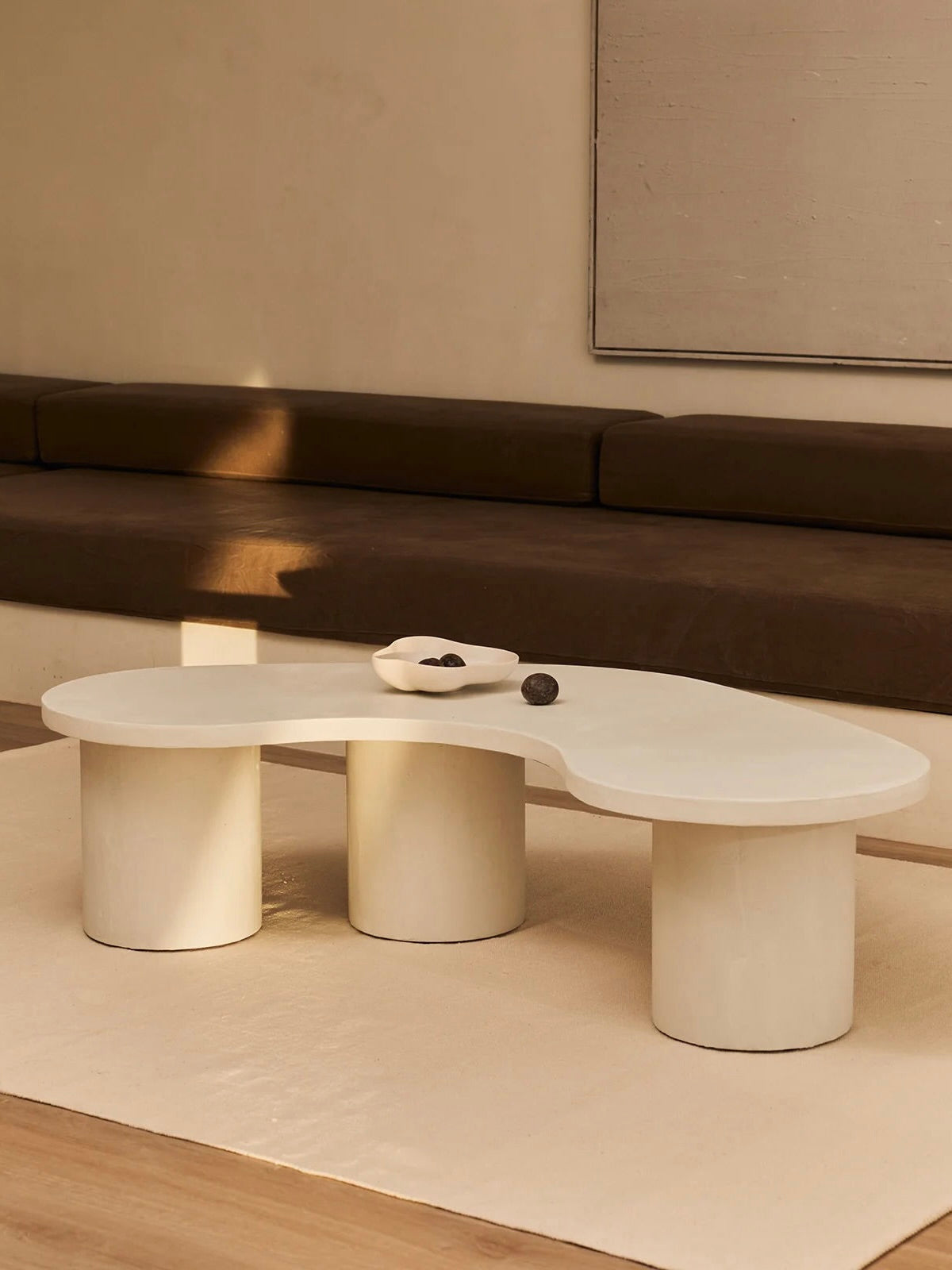In a minimalist living room, MarlotBaus's "Parmigiano" Coffee Table boasts an abstract wavy shape with cylindrical legs. A small white dish with decorative stones sits on the table, while a beige couch with brown cushions completes the sophisticated look against a neutral microcement wall.
