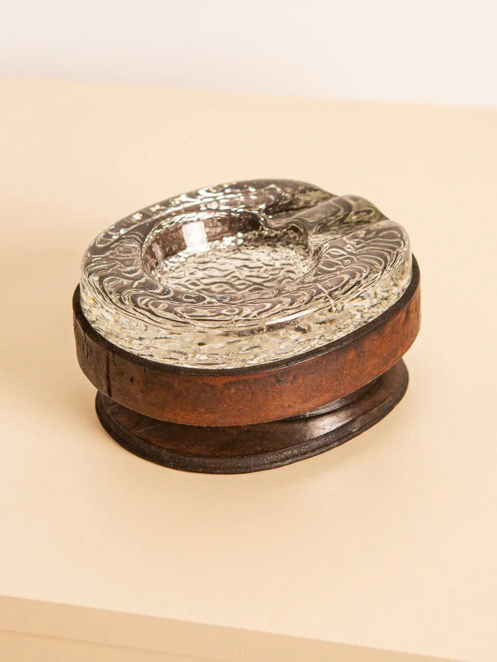 The Treaptyque Murano Glass Ashtray With Leather, featuring intricate designs, rests elegantly on a round, rustic wooden base atop a light-colored surface, exuding Italian design and retro charm.