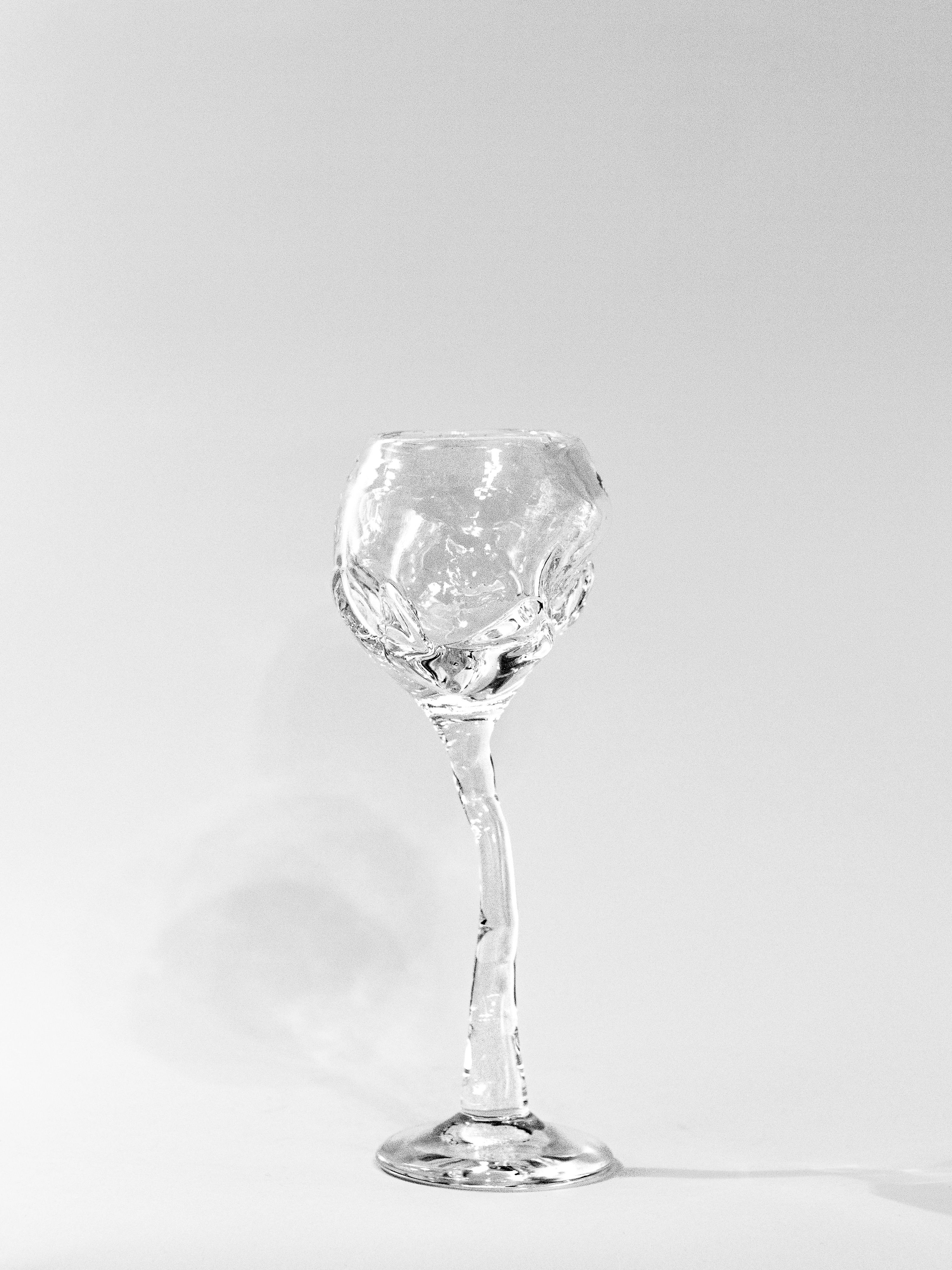 The Hand Blown Freeform Wine Glass by Szkło Studio features a distinctive shape with a textured surface and uniquely curved stem, crafted from transparent sodium glass. It is set against a plain white background with shadows cast to the left.