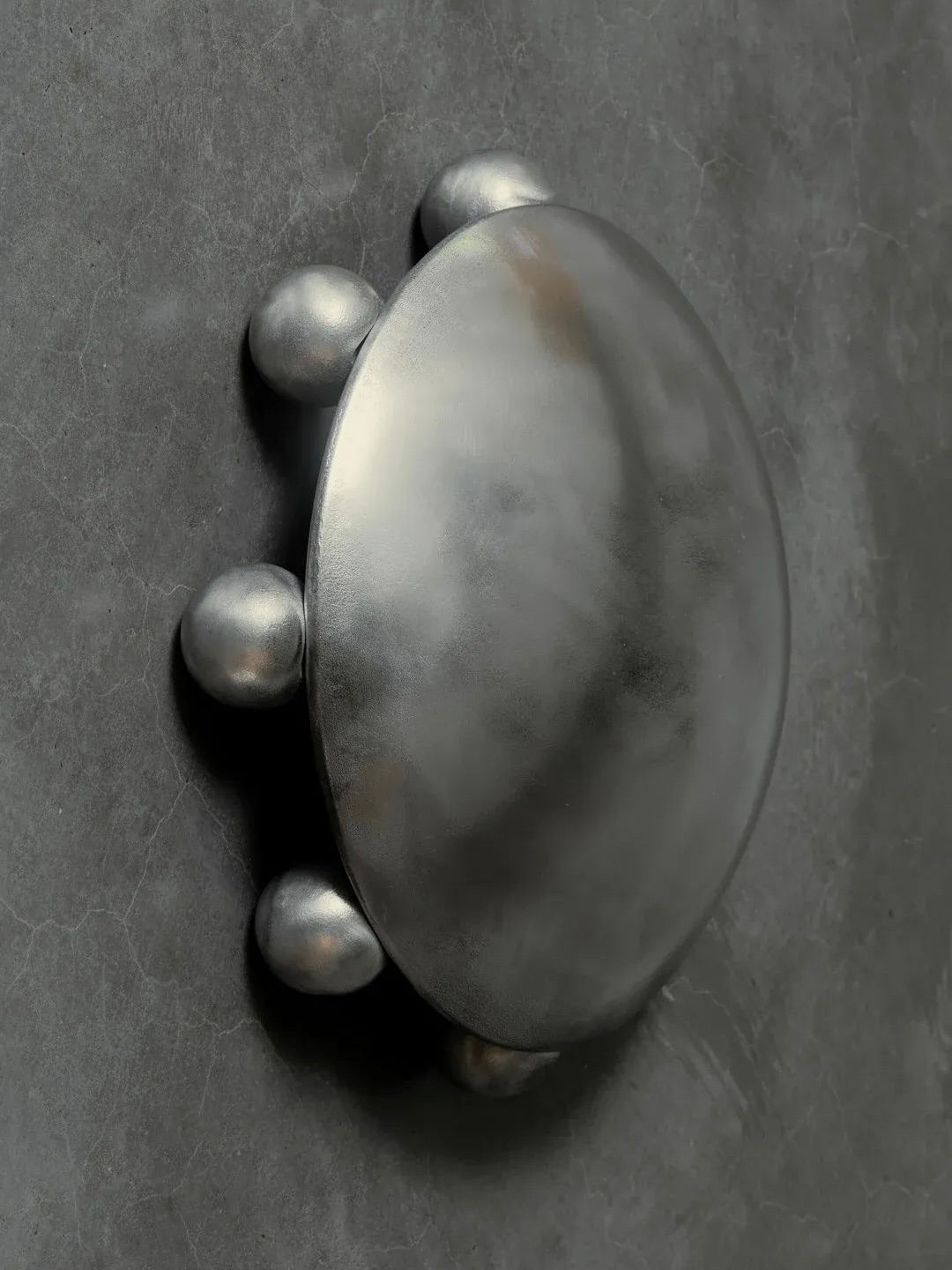 The "ALTER" Sconce by Kaytar captures the art of transformation with its circular silver design, four round protrusions, and intricately sand-cast textured gray surface, resembling a futuristic decorative piece showcasing the essence of alchemy.