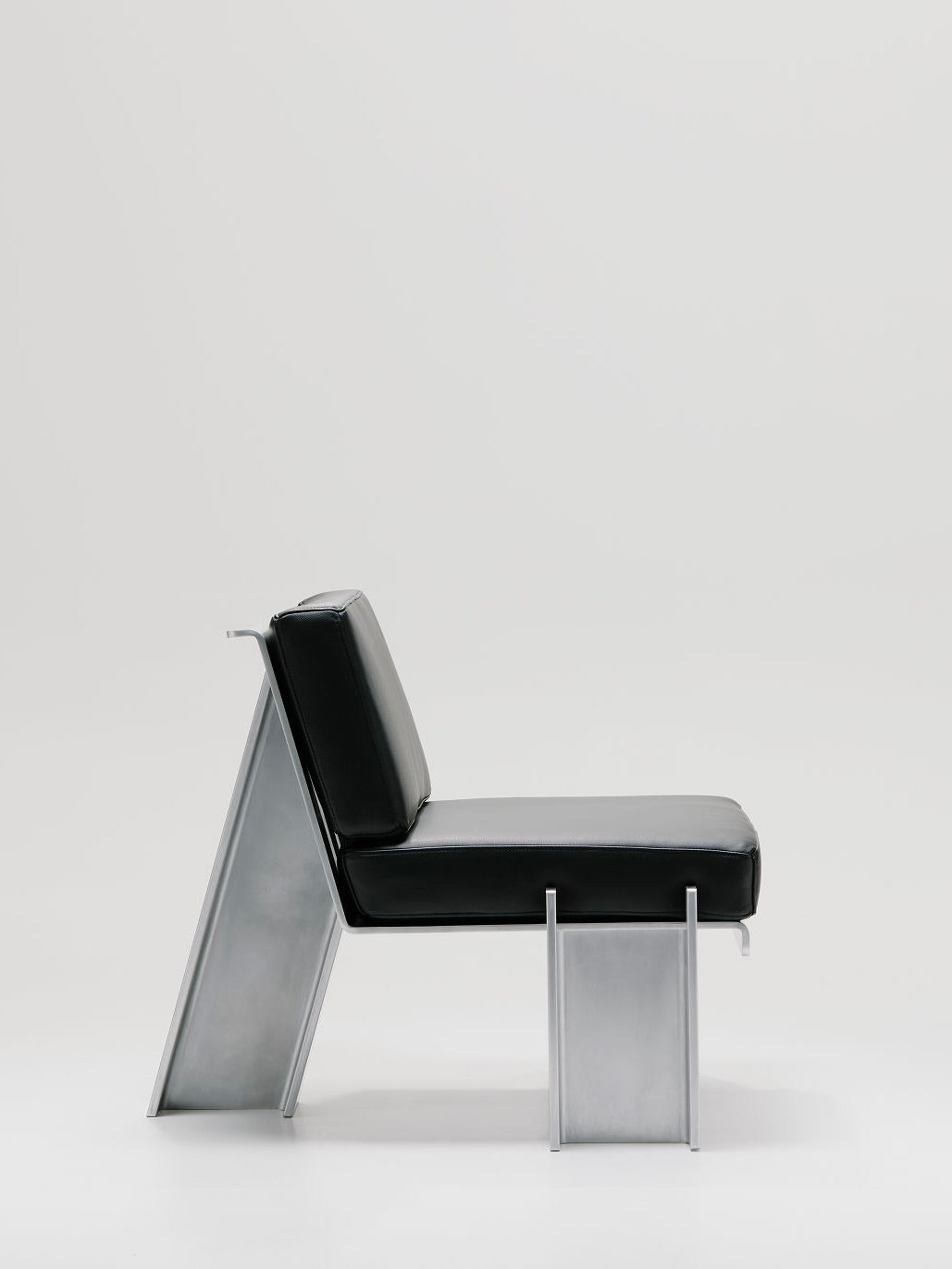 Side view of the Marquel Williams Beam Lounge Chair, showcasing a sleek black cushion and geometric waxed aluminum frame, on a white background, highlighting its angular supports and minimalist design.