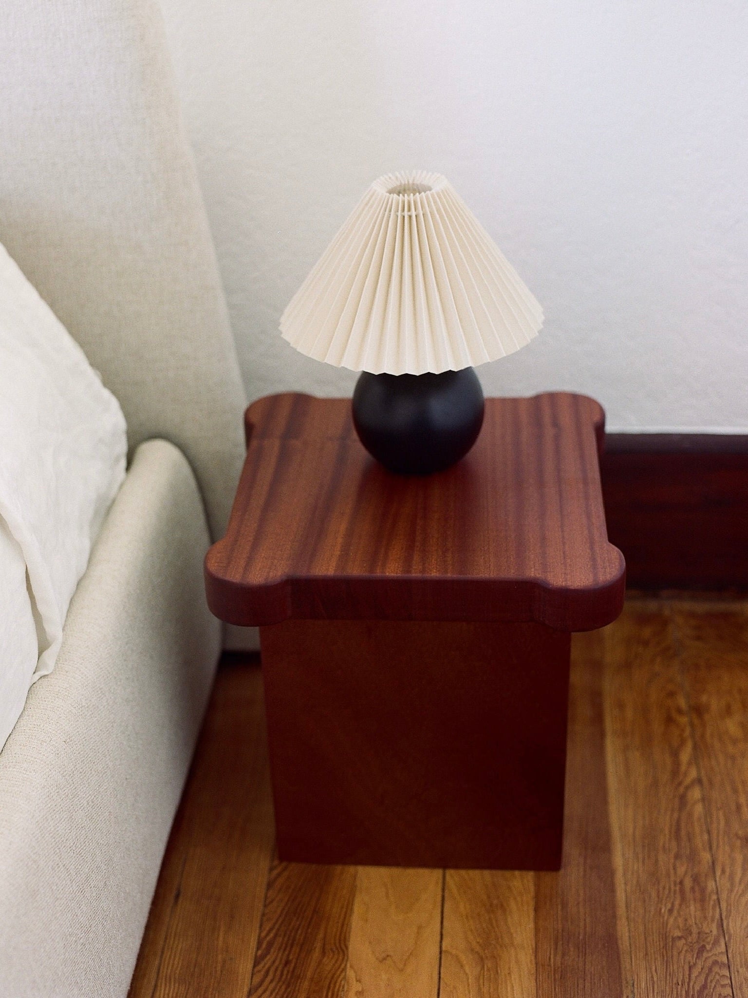 A small, dark wooden bedside table with a wavy edge from MUHLY, known as the Bijou Mini Table, sits on a wooden floor beside a light-colored bed. A black lamp with a pleated white shade is placed on the table. The white wall behind and minimalist design complete the serene space.