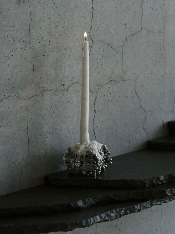 A tall lit white candle with melted wax sits in a Panorammma Chrome Candle Holder on a dark, textured stone ledge against a cracked concrete wall, creating a moody, atmospheric scene reminiscent of organic forms.