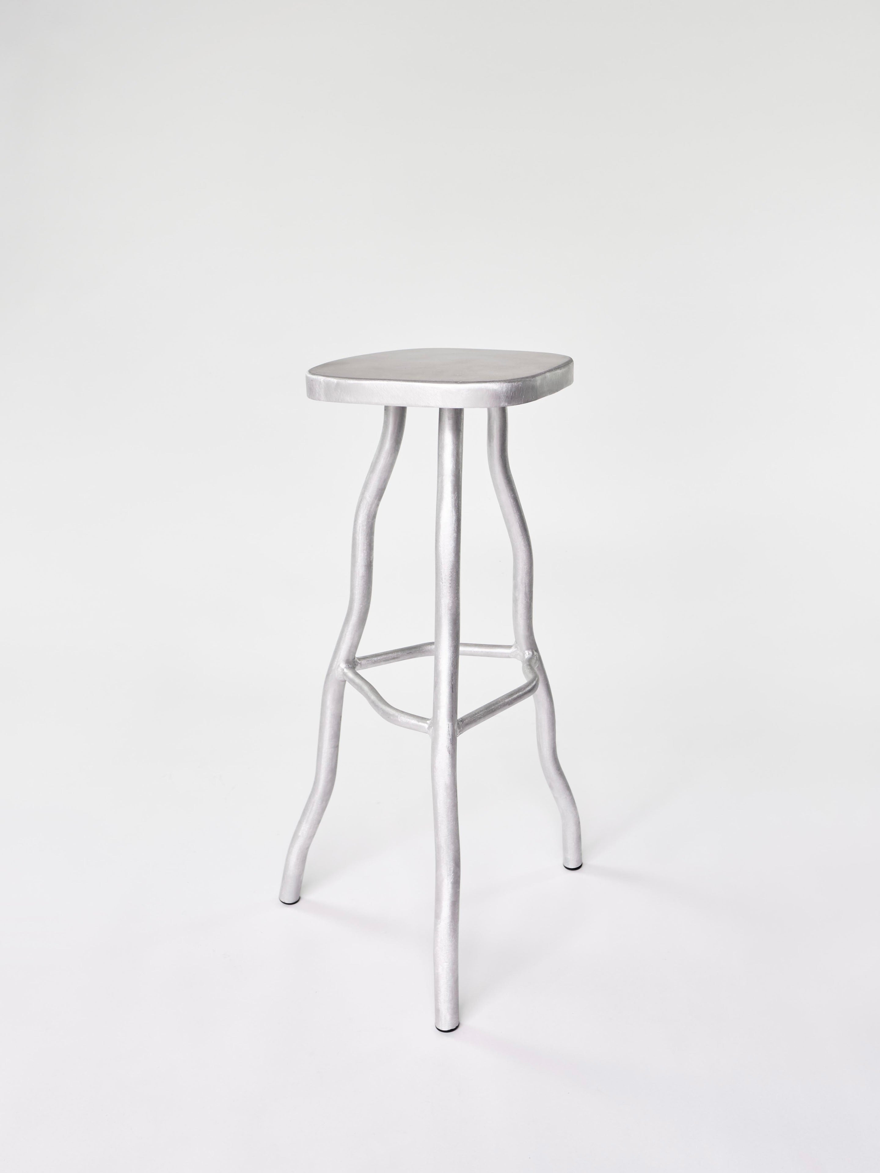 A tall, handcrafted silver metal bar stool by Six Dots Design features a minimalist design with a round seat and four slender, outward-curving legs connected by horizontal supports. Set against a plain white background, it exudes elegance and simplicity.