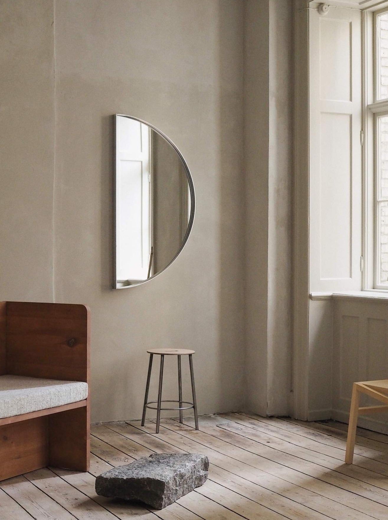 A serene room with a wooden floor and minimalistic decor, featuring a FRAMA recess mirror on the wall, a window with natural light, a simple bed, a stool, and a unique rock-like object
