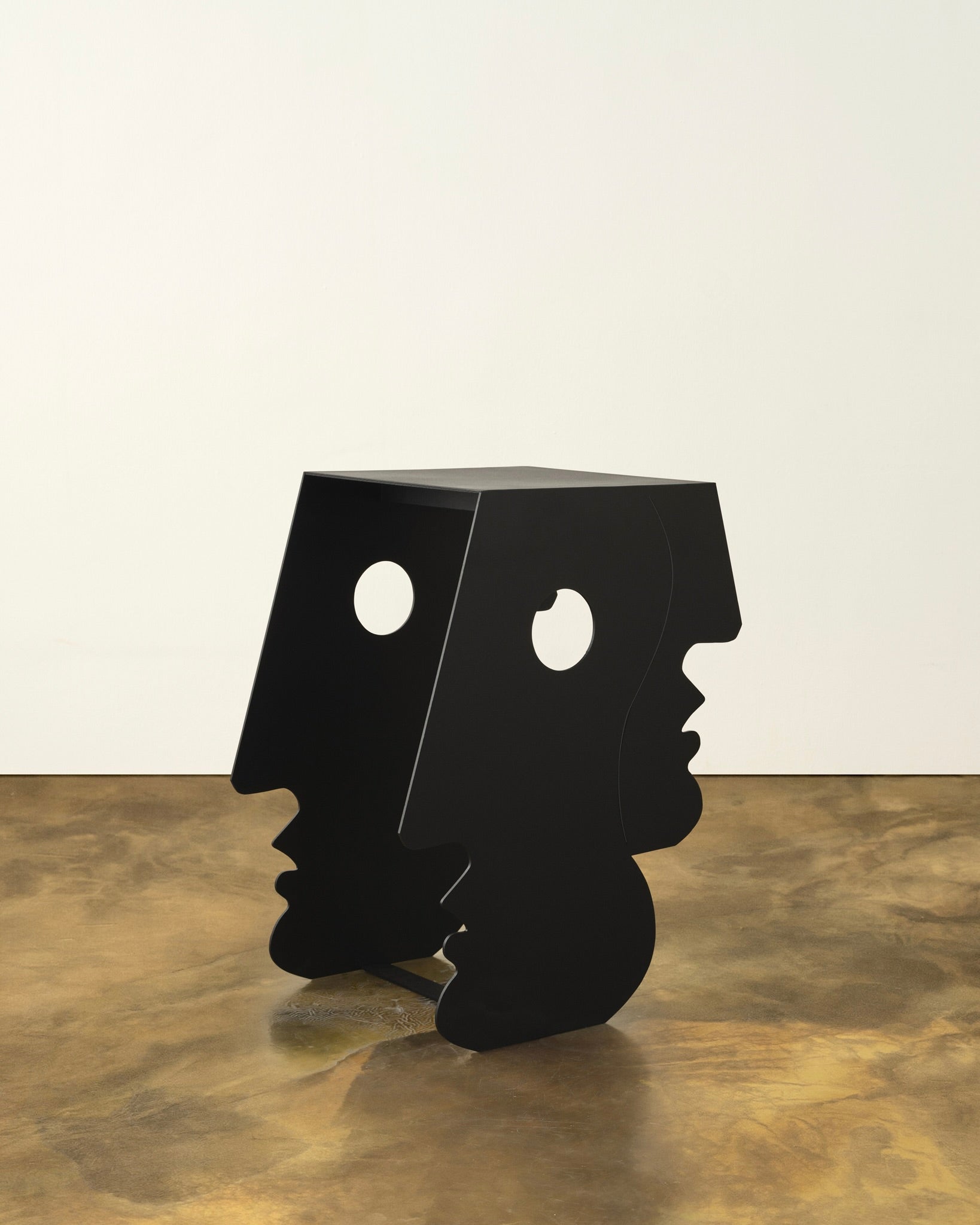 The "Lucia" Side Table in Black by Animate Objects features Bauhaus-inspired sides shaped like stylized human profiles with circular eye cut-outs, resting on a polished concrete floor against a plain light wall—ideal as a unique accent piece.