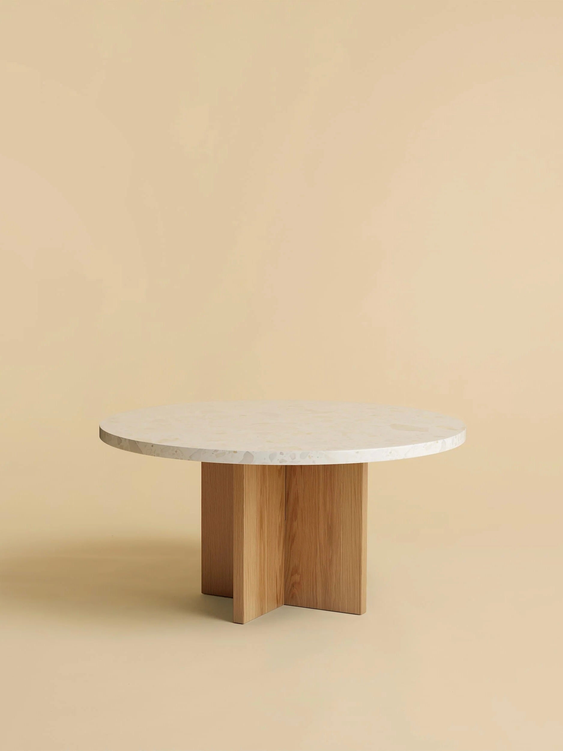 The "Tinian" Coffee Table by Lebanto, showcasing an artisan-crafted smooth white marble top and a wooden cross-shaped base, is set against a beige background.