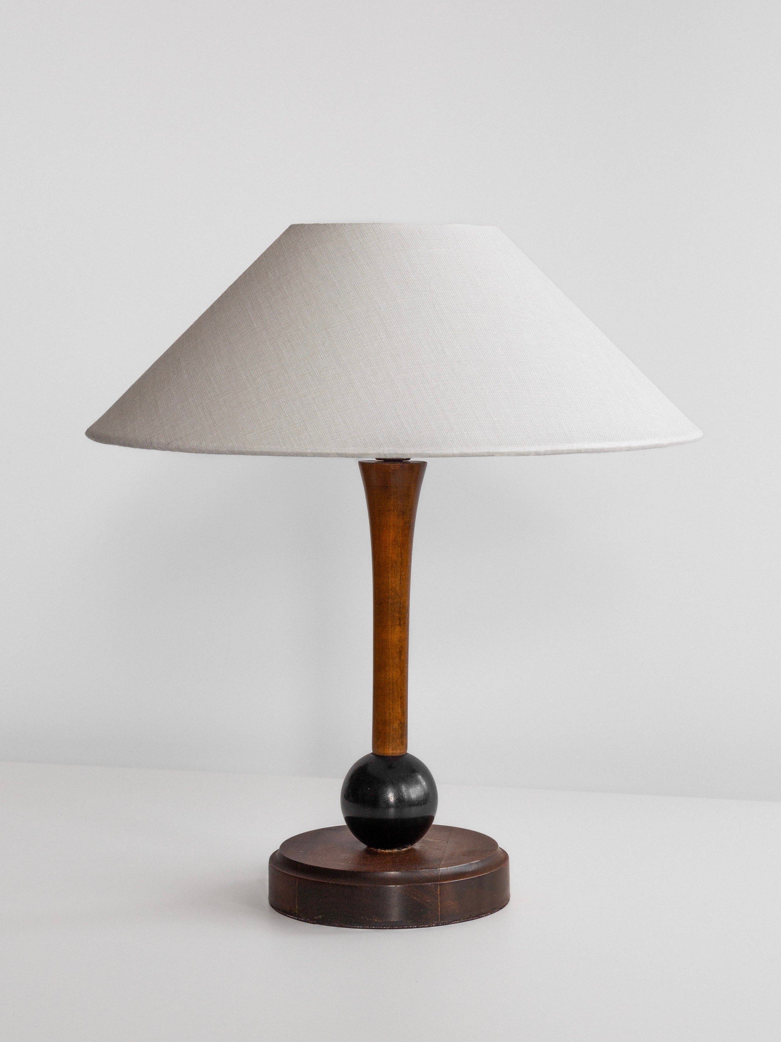 Introducing the Wooden Art Deco Table Lamp by Spigel: a 1930s Sweden-inspired lamp featuring a wide, conical white lampshade and a sleek wooden base with a round black accent. The minimalistic and elegant design is reminiscent of the Art Deco style, perfect against a plain, light-colored background.