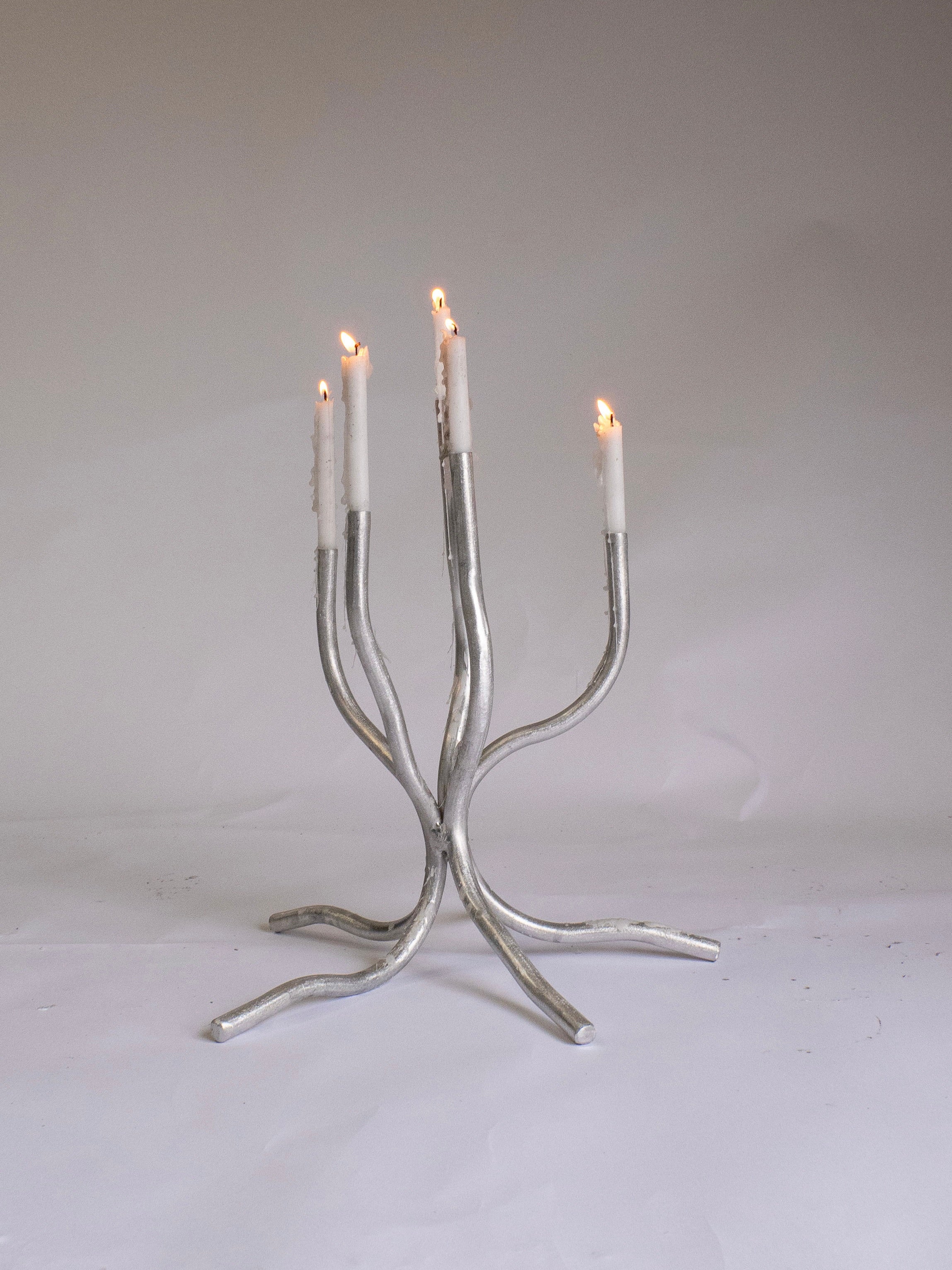 A Small Candelabra by Six Dots Design, made of silver metal with four branches each holding a lit white candle, stands on a plain white surface against a light gray background.