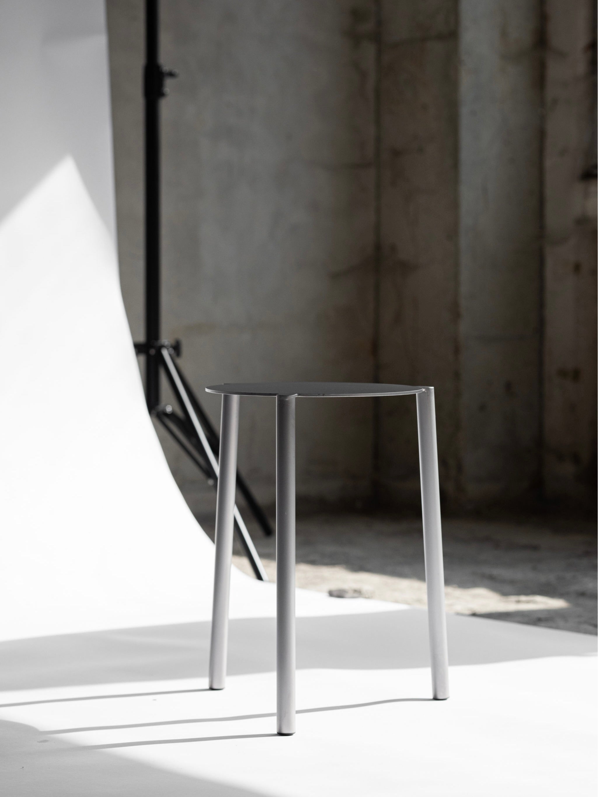 The Tabouret Isla x Paul Delrez from KØGE Design, evoking the seating comfort of Parisian bistros, showcases a minimalist aesthetic with three slim legs. Set against a white backdrop, it is illuminated by natural side light, creating a gentle shadow on the floor.