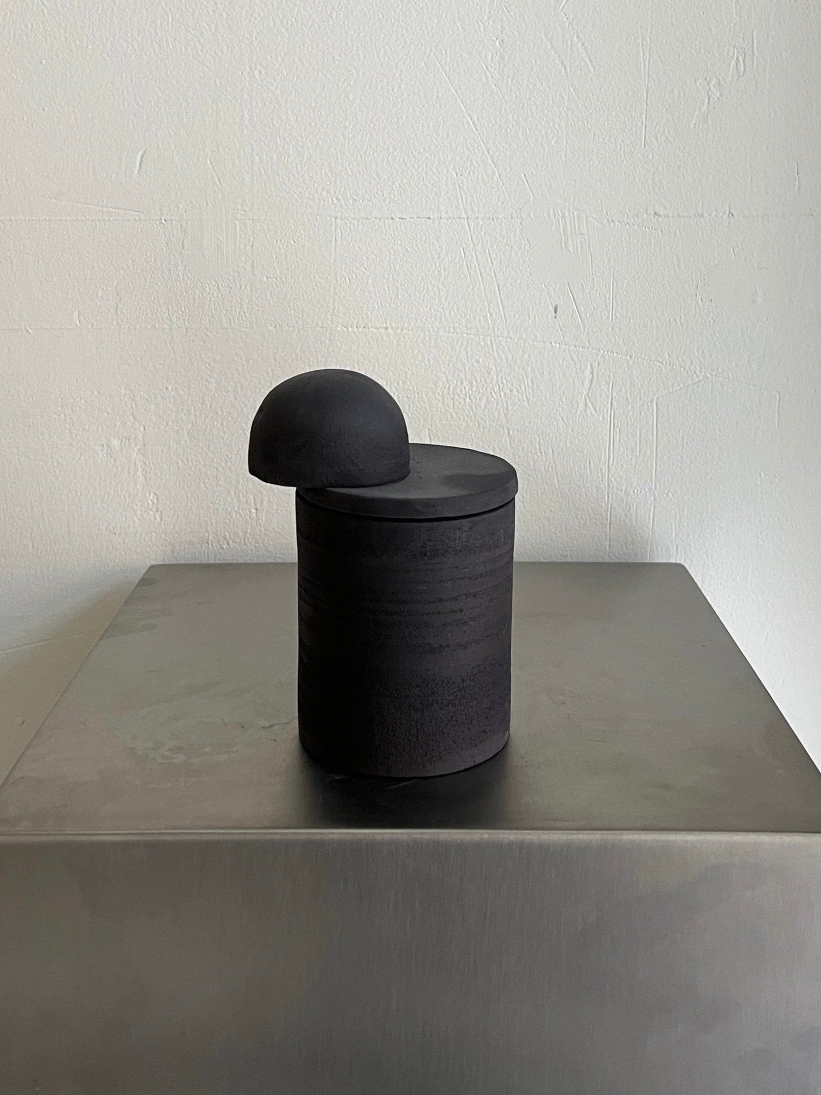 The Sculptural Scented Candle no.8 Black by 2222Studio, a sleek minimalist black ceramic container with a half-sphere lid, rests on a metallic surface. This limited edition fills the room with earthy patchouli, fir needle, and blood orange scents against a plain light-colored wall background.
