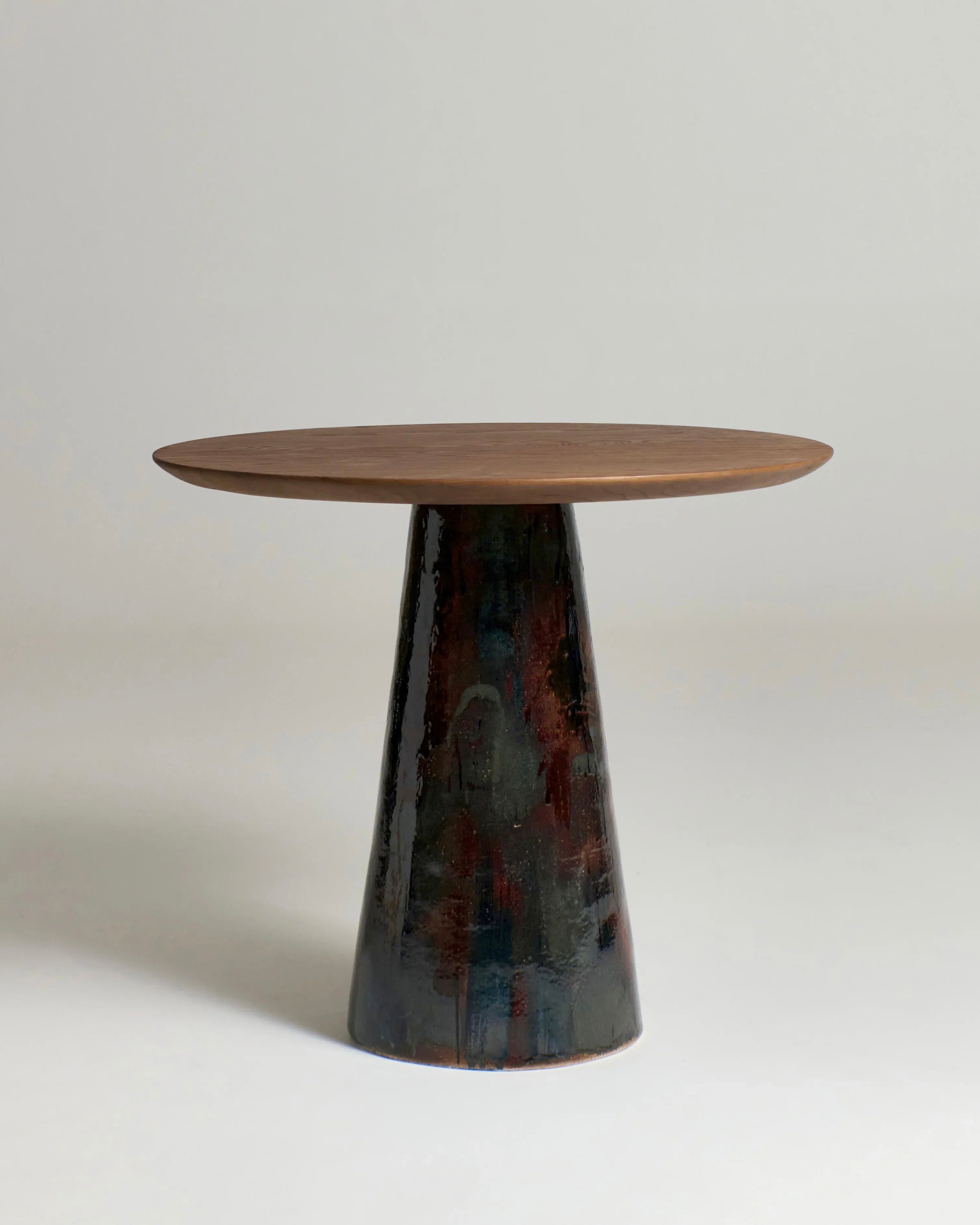 The Porto Belo Table by Project 213A features a round wooden surface with a rustic finish, elegantly supported by a wide, tapered, hand-sculpted ceramic base that adds texture and intrigue against its plain, light-colored backdrop.