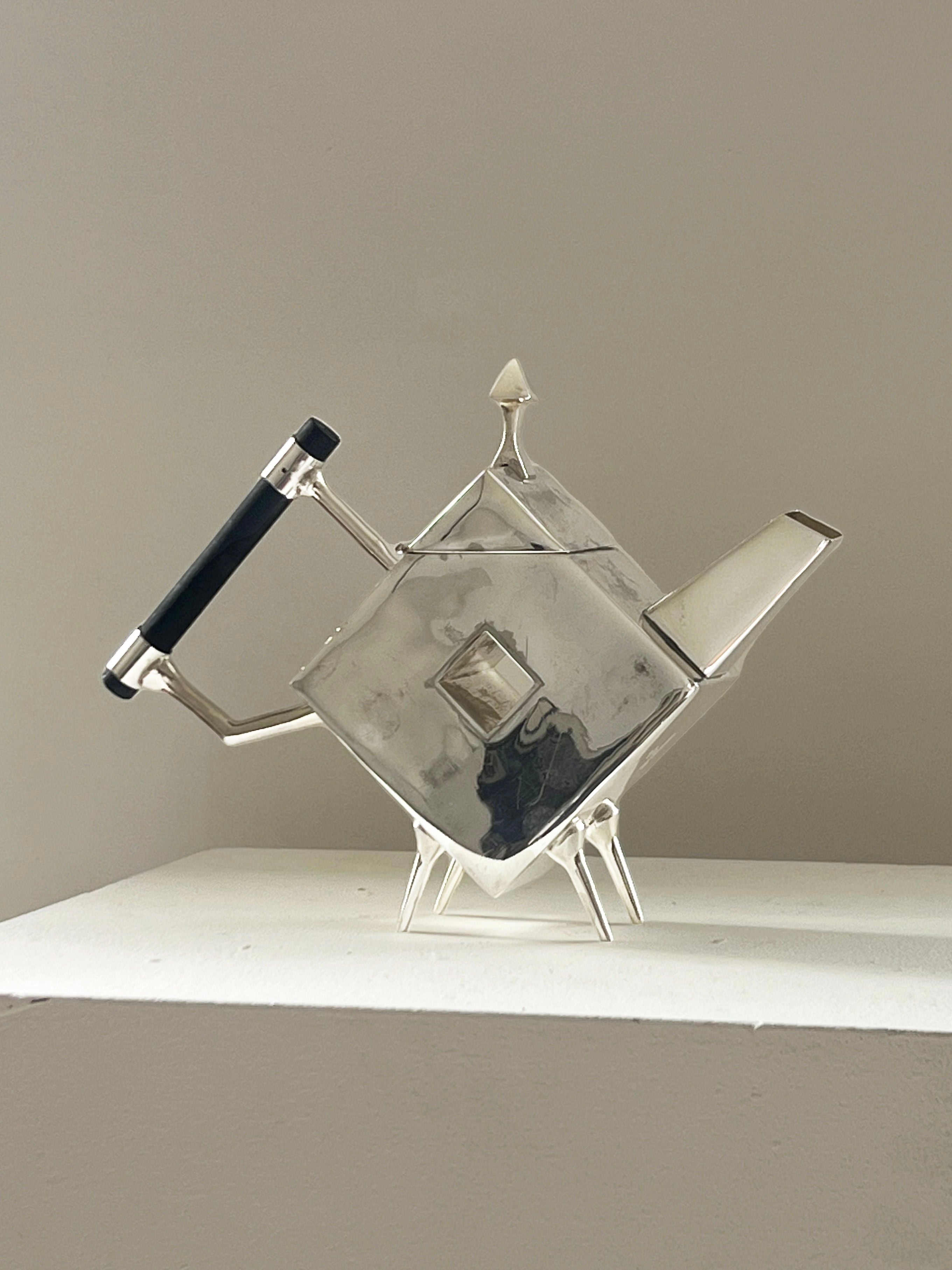 The Christopher Dresser Square Teapot by Porter Studio is modern and angular, silver-plated with a black handle, featuring sharp edges and a triangular spout. It sits on a white surface against a neutral background, showcasing its unique geometric design.