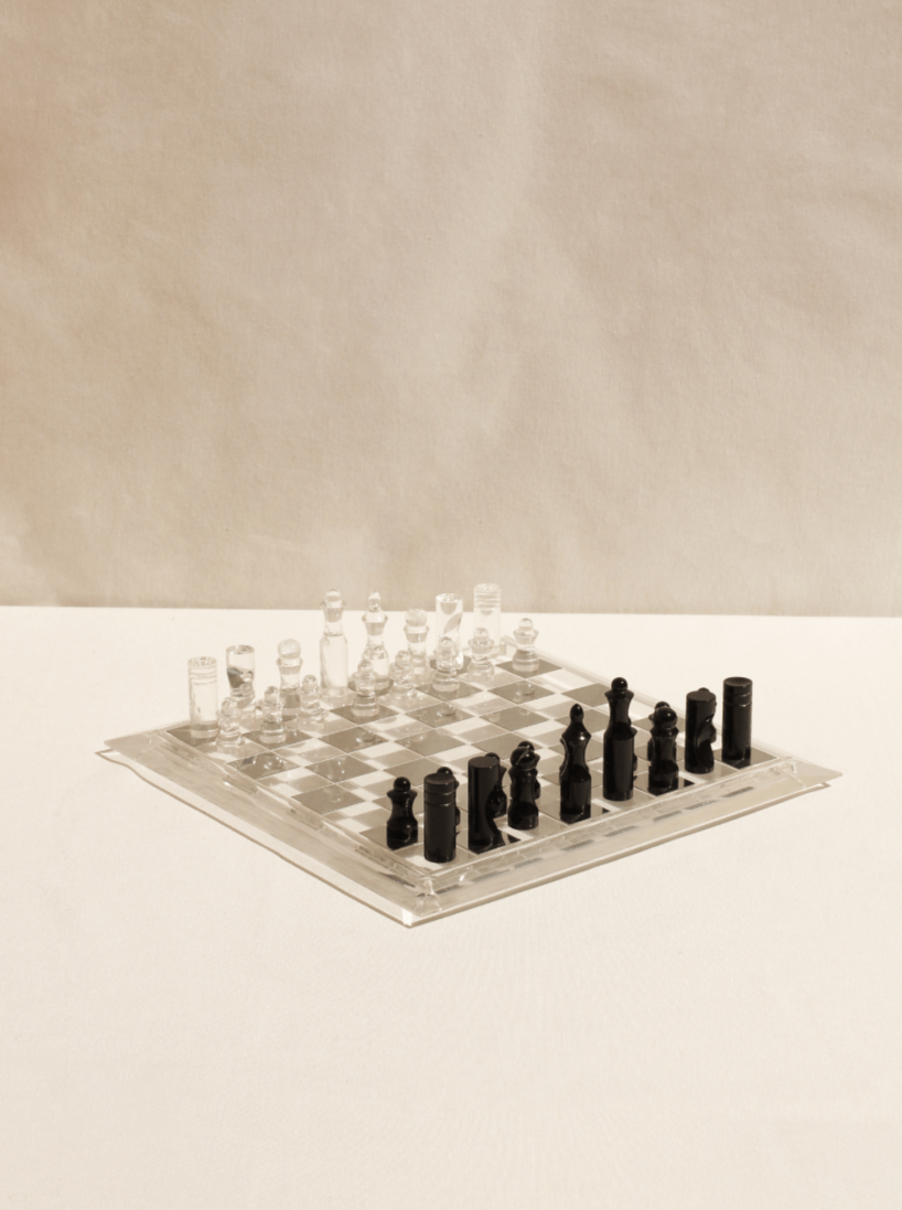 The Boga Avante Shop's Acrylic Chess Board, ideal for luxury interiors, features clear and black glass pieces arranged for a new game on a light surface. This sculptural set softly reflects light, embodying minimalist aesthetics.